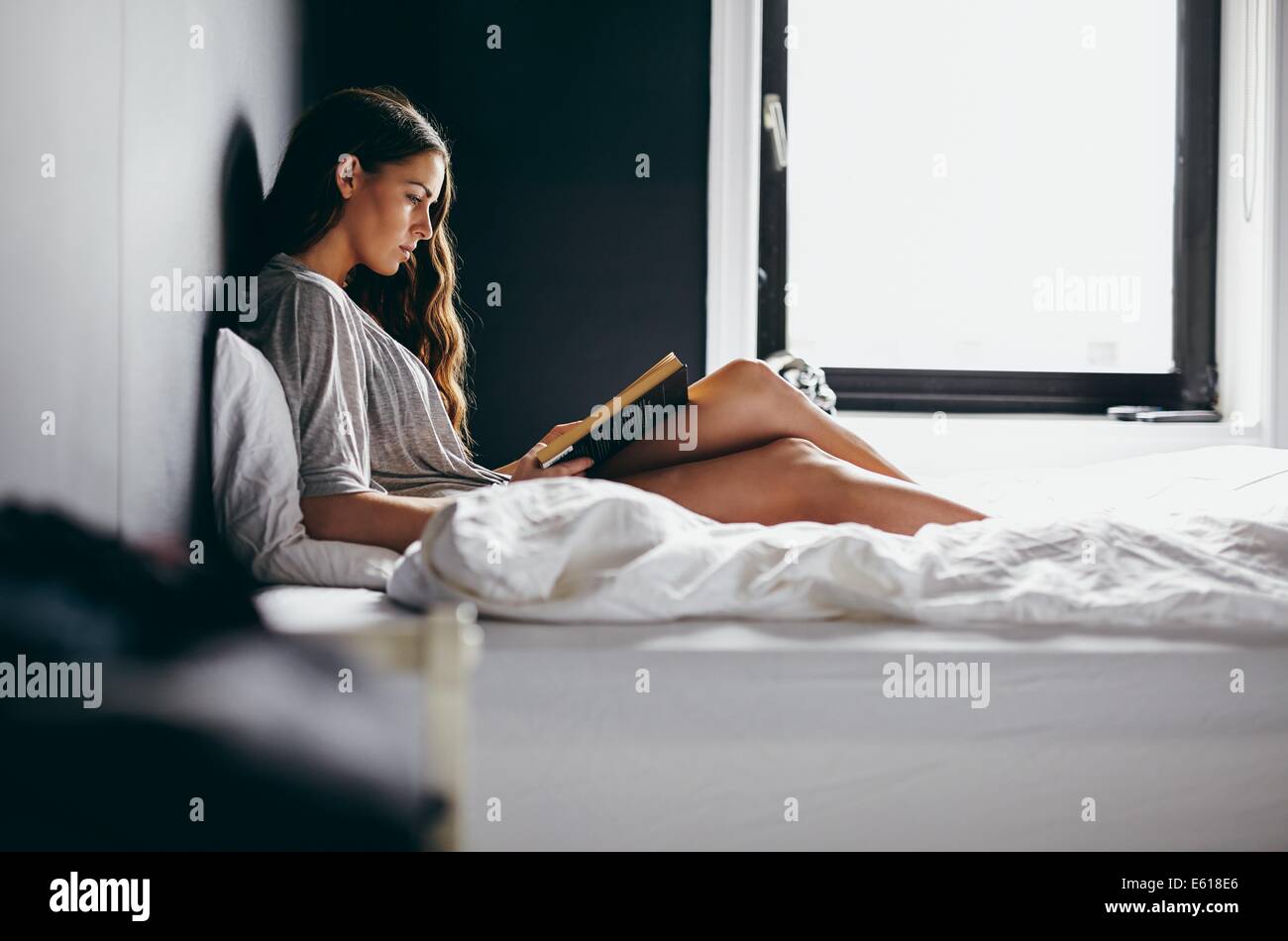 Vue latérale d'une grenaille belle jeune femme assise sur son lit la lecture d'un roman intéressant. Modèle des femmes de race blanche dans la chambre. Banque D'Images