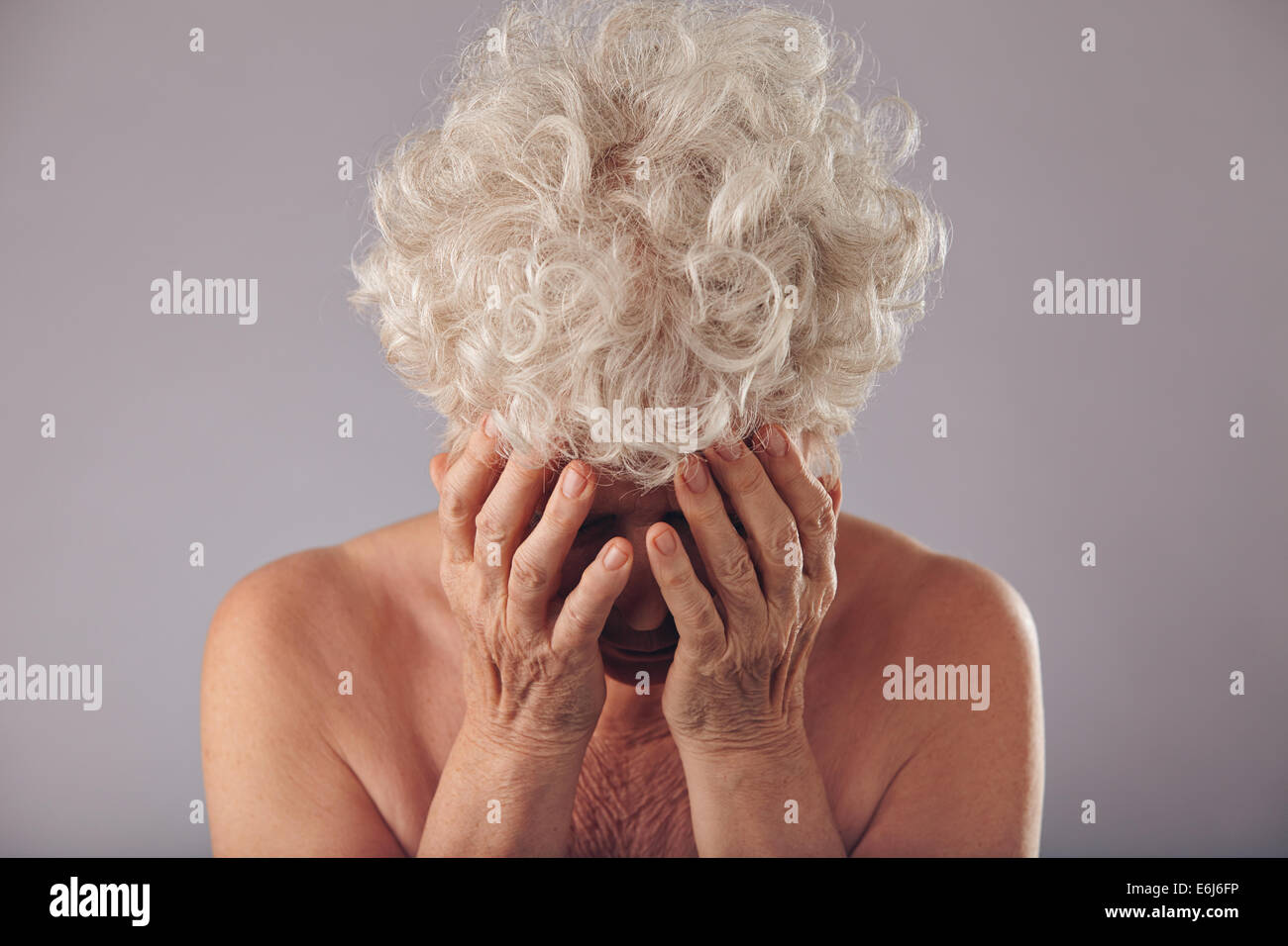 Portrait de vieille dame malheureuse holding il visage dans les mains à la recherche vers le bas, contre un arrière-plan gris. Sad senior woman. Banque D'Images
