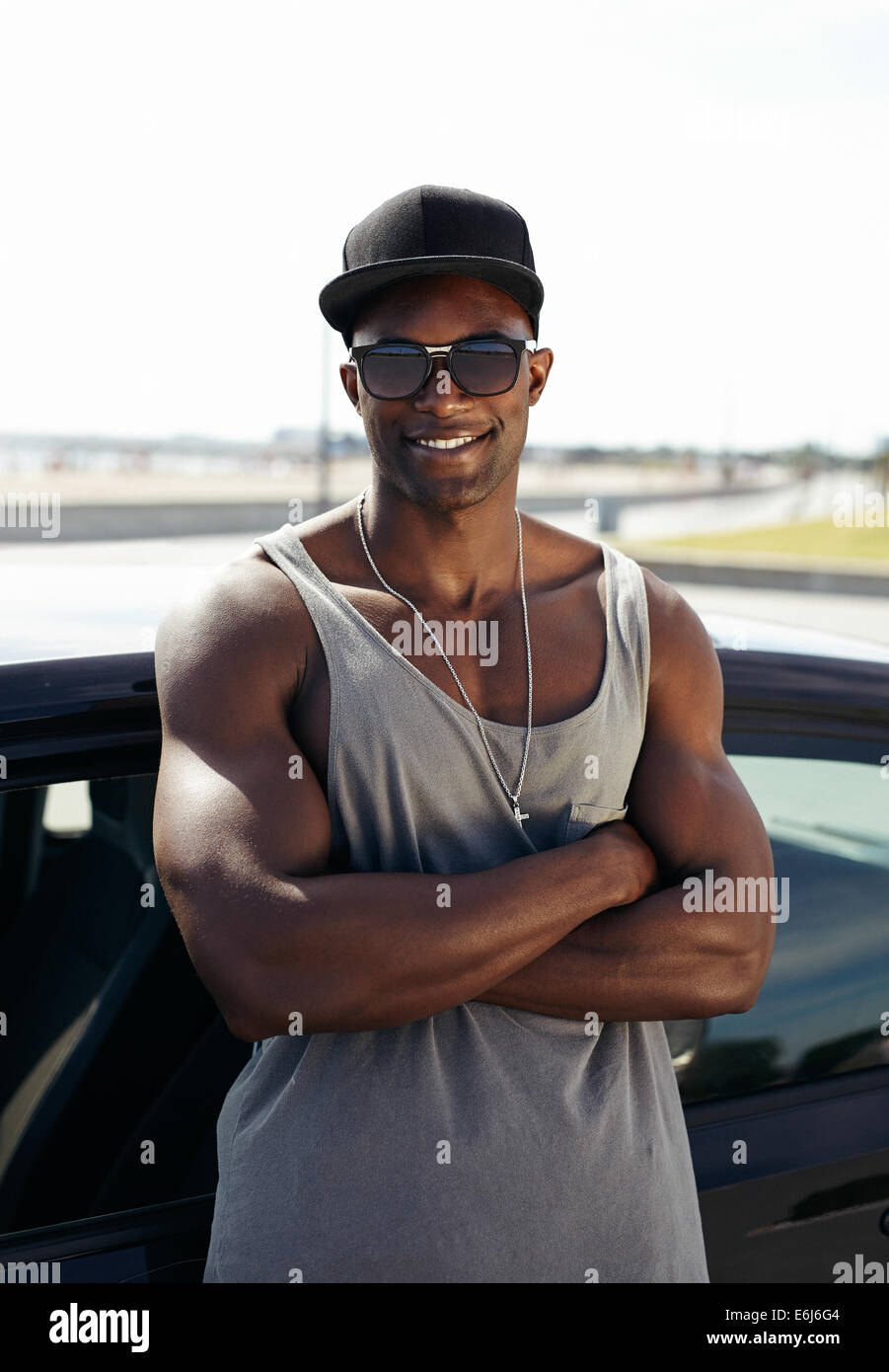 Portrait of handsome guy africains avec les bras croisés appuyé contre une voiture en souriant. Guy musculaire stylisé portant des lunettes de soleil. Banque D'Images