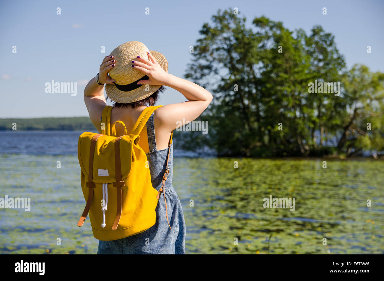 Une jeune femme avec un sac à dos jaune tenant son chapeau devant un lac et  arbres en été Photo Stock - Alamy