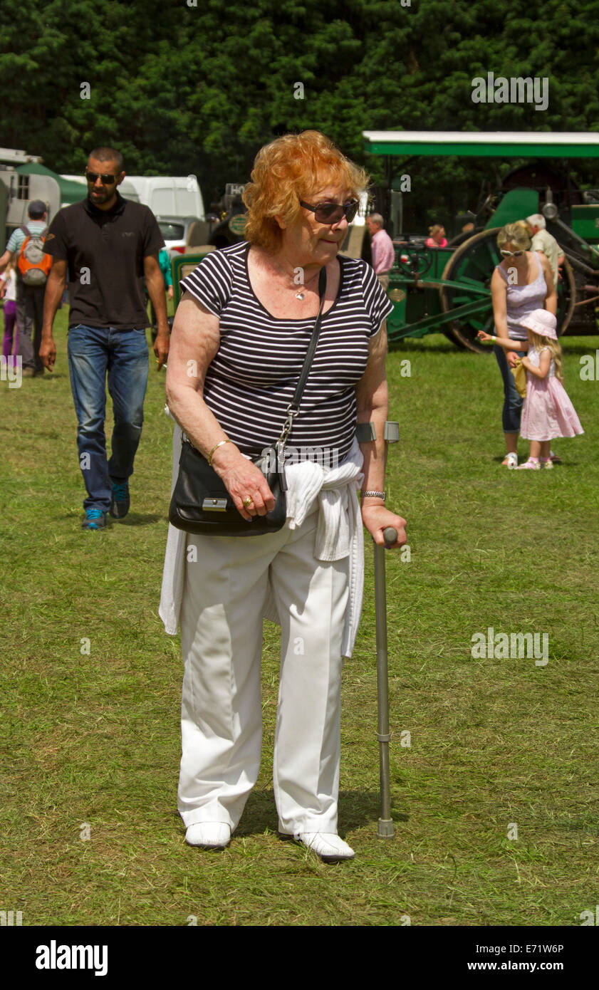 Vieille Femme aux cheveux rouges et portant des T-shirt à rayures et un pantalon blanc à marcher avec l'aide d'un bâton Banque D'Images