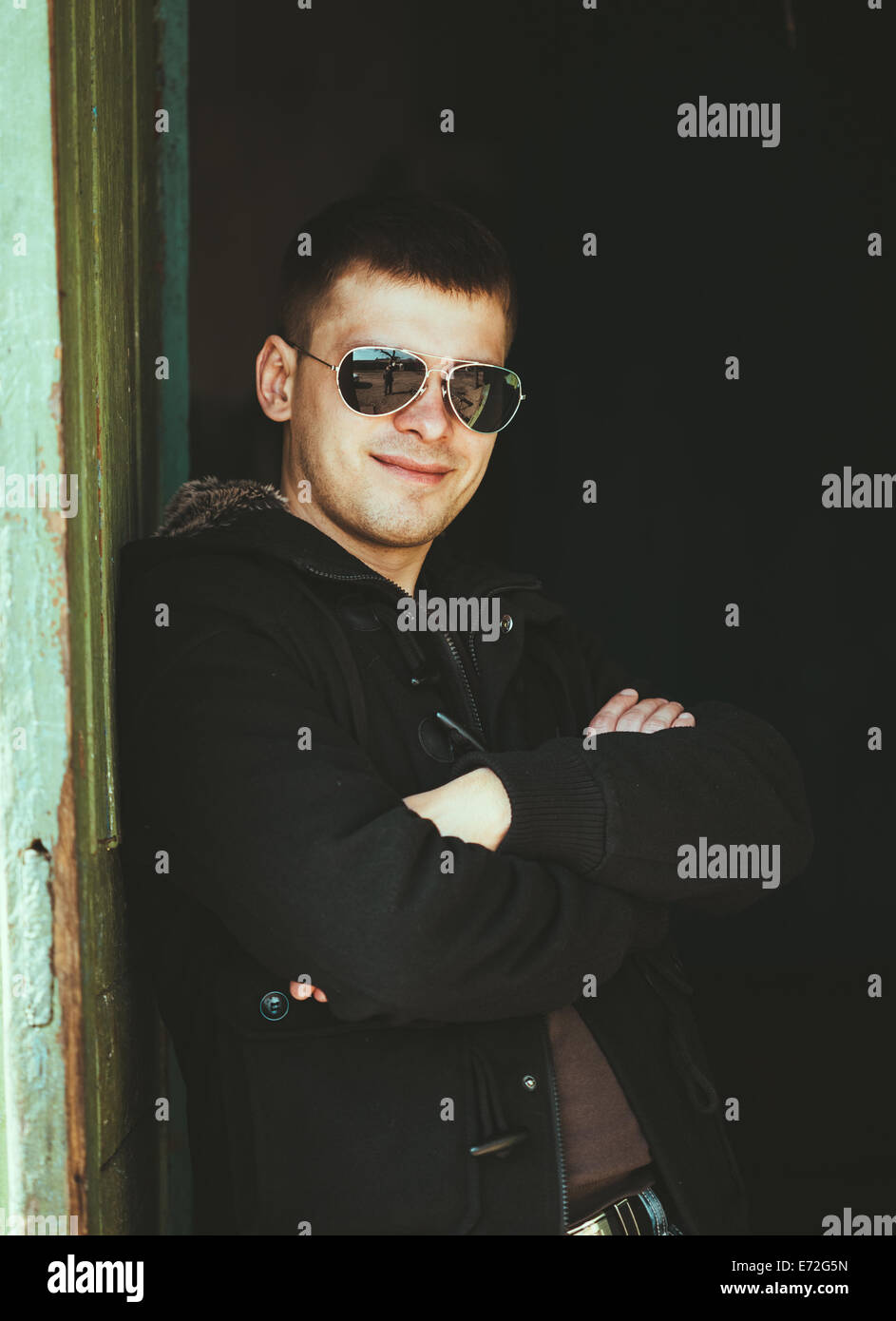 Beau jeune homme d'un séjour près de vieille maison de bois dans la journée d'automne, de détente, de réflexion, de sourire. Style Casual - veste, lunettes de soleil. Banque D'Images