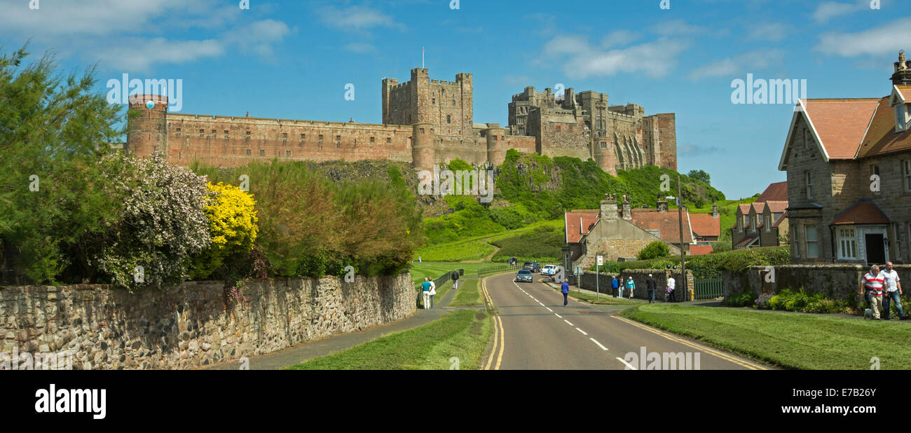 Vue panoramique de l'immense et historique château de Bamburgh sur colline herbeuse avec des chalets de la ville et les gens en premier plan dans le cadre de ciel bleu en Angleterre Banque D'Images