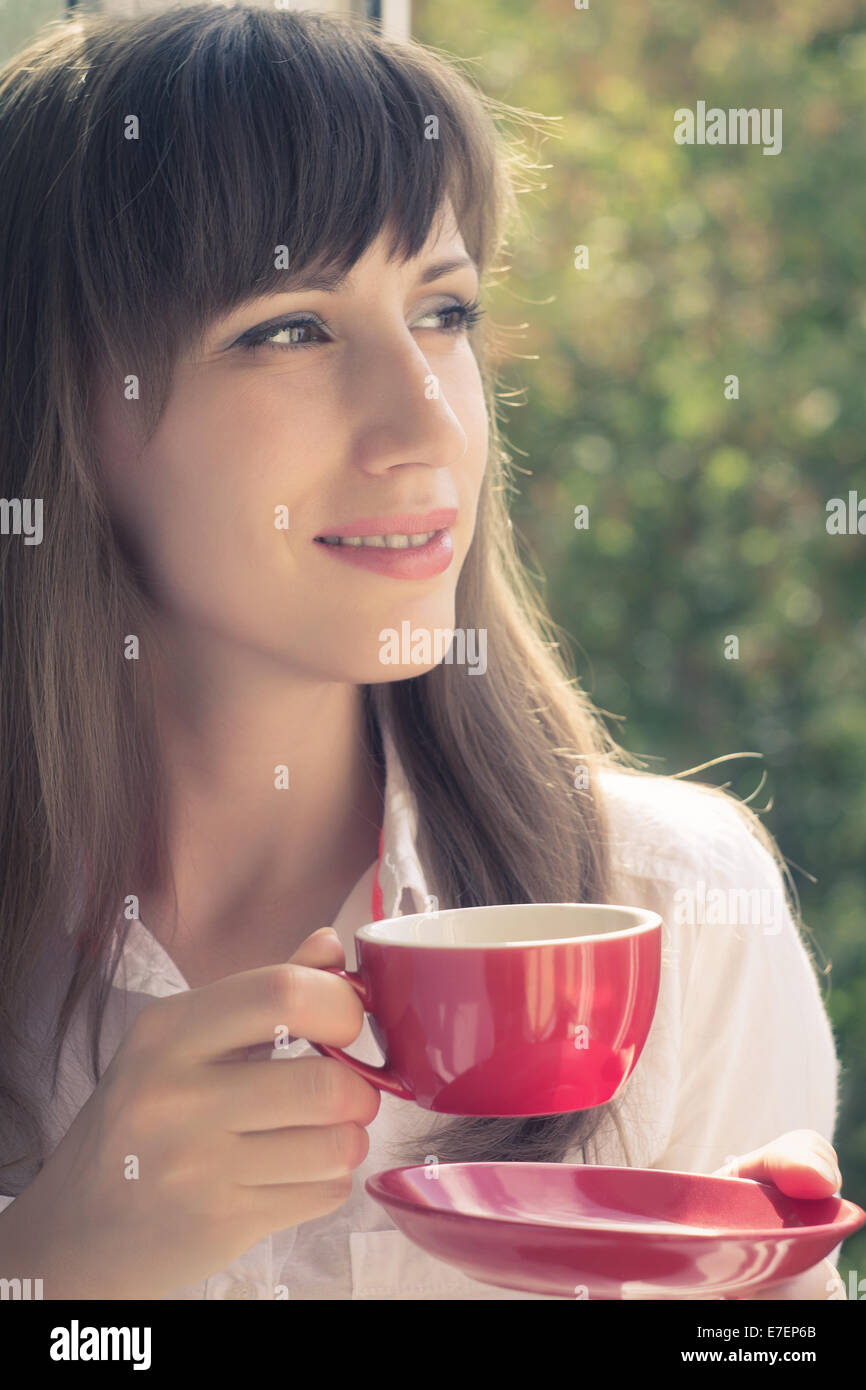 Belle jeune femme hipster boire le thé près de la fenêtre. Beauté caucasien fille avec tasse de café en regardant par la fenêtre. Banque D'Images