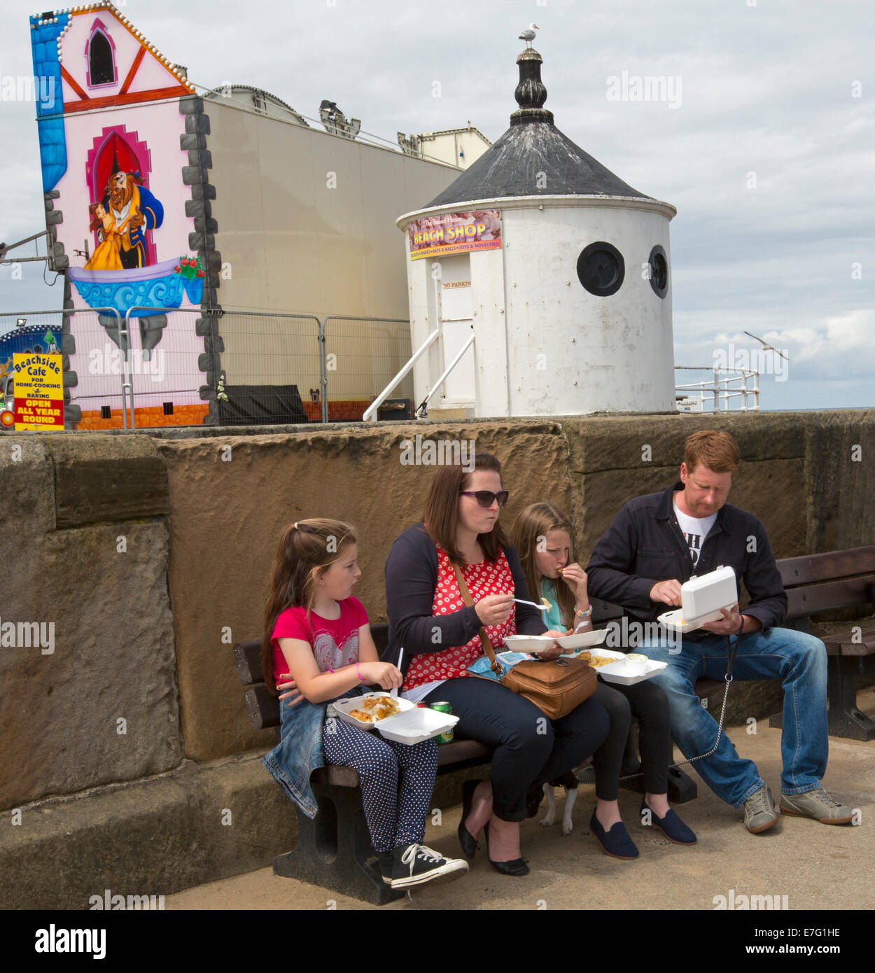 Famille avec deux enfants assis sur le mur de manger le déjeuner de poisson et frites de conteneurs à takeway English ville de bord - Whitby Banque D'Images