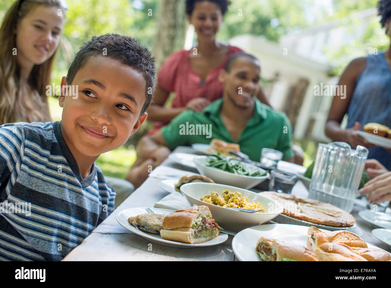 Une réunion de famille, hommes, femmes et enfants autour d'une table dans un jardin en été. Un garçon souriant au premier plan. Banque D'Images