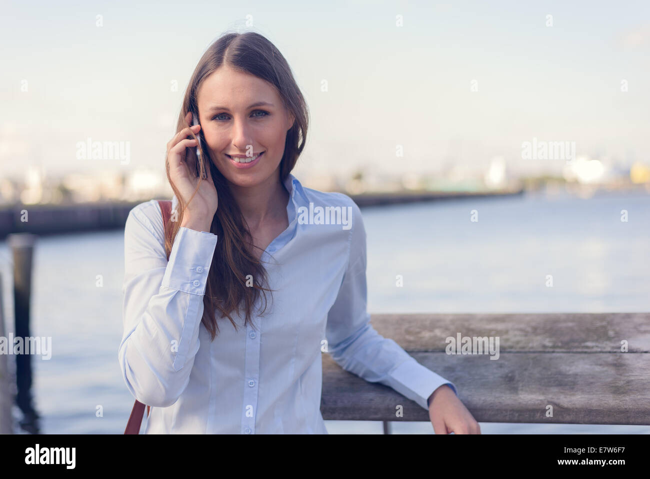 Jolie femme de prendre un appel sur son téléphone portable à l'extérieur debout contre un arrière-plan de la rivière smiling at the camera Banque D'Images
