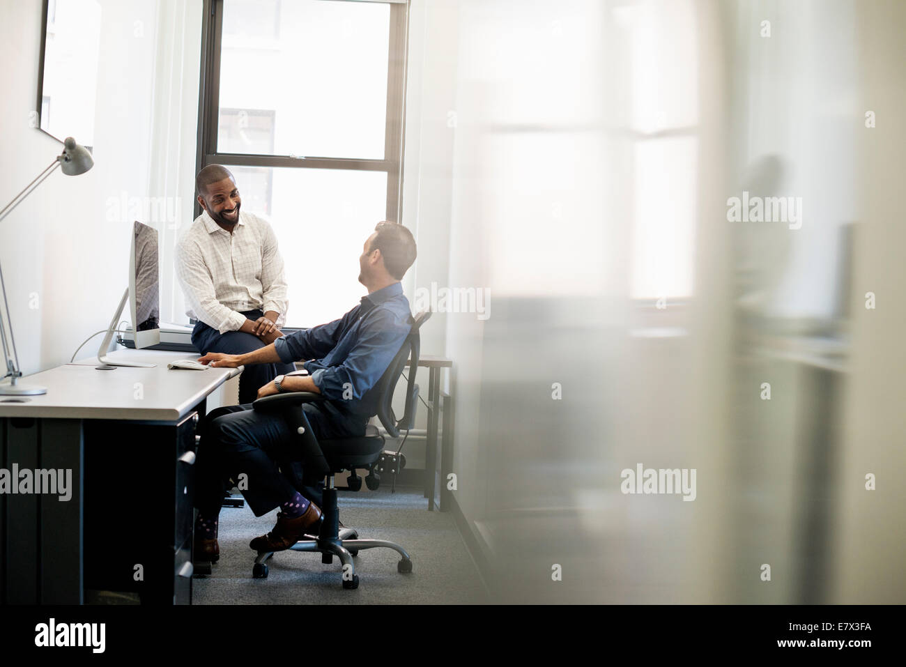 La vie de bureau. Un homme penché en arrière dans une chaise de bureau parlant à un collègue assis sur le bord du bureau. Banque D'Images