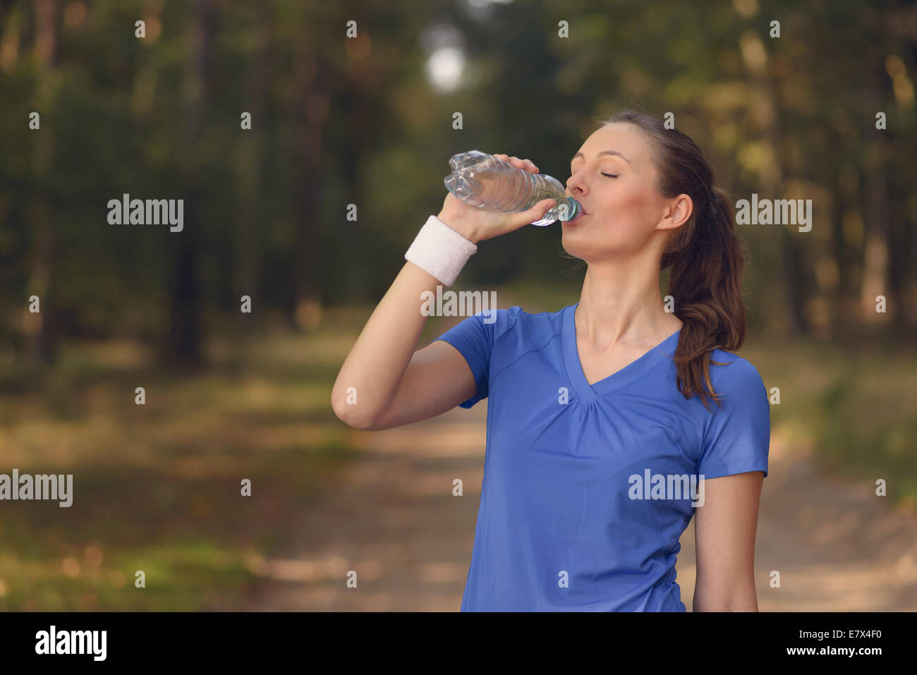 Jeune femme élancée fit boire de l'eau embouteillée dans les vêtements de sport alors qu'elle s'arrête sur une piste forestière à réhydrater pendant un run de formation Banque D'Images