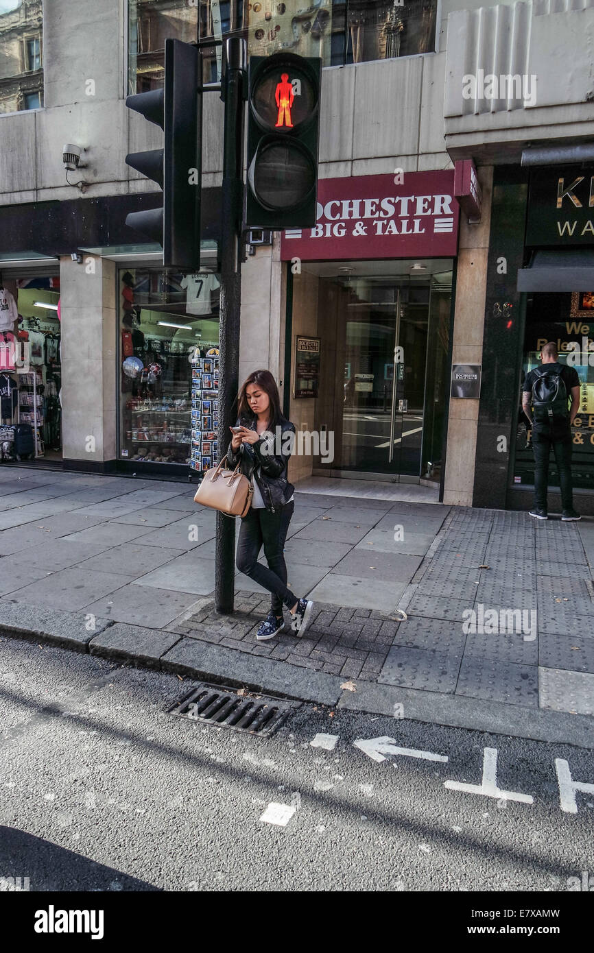 Une jeune fille s'appuie contre un feu de circulation à l'aide de son téléphone portable tenant un sac à main dans l'attente de traverser la route à Londres Banque D'Images