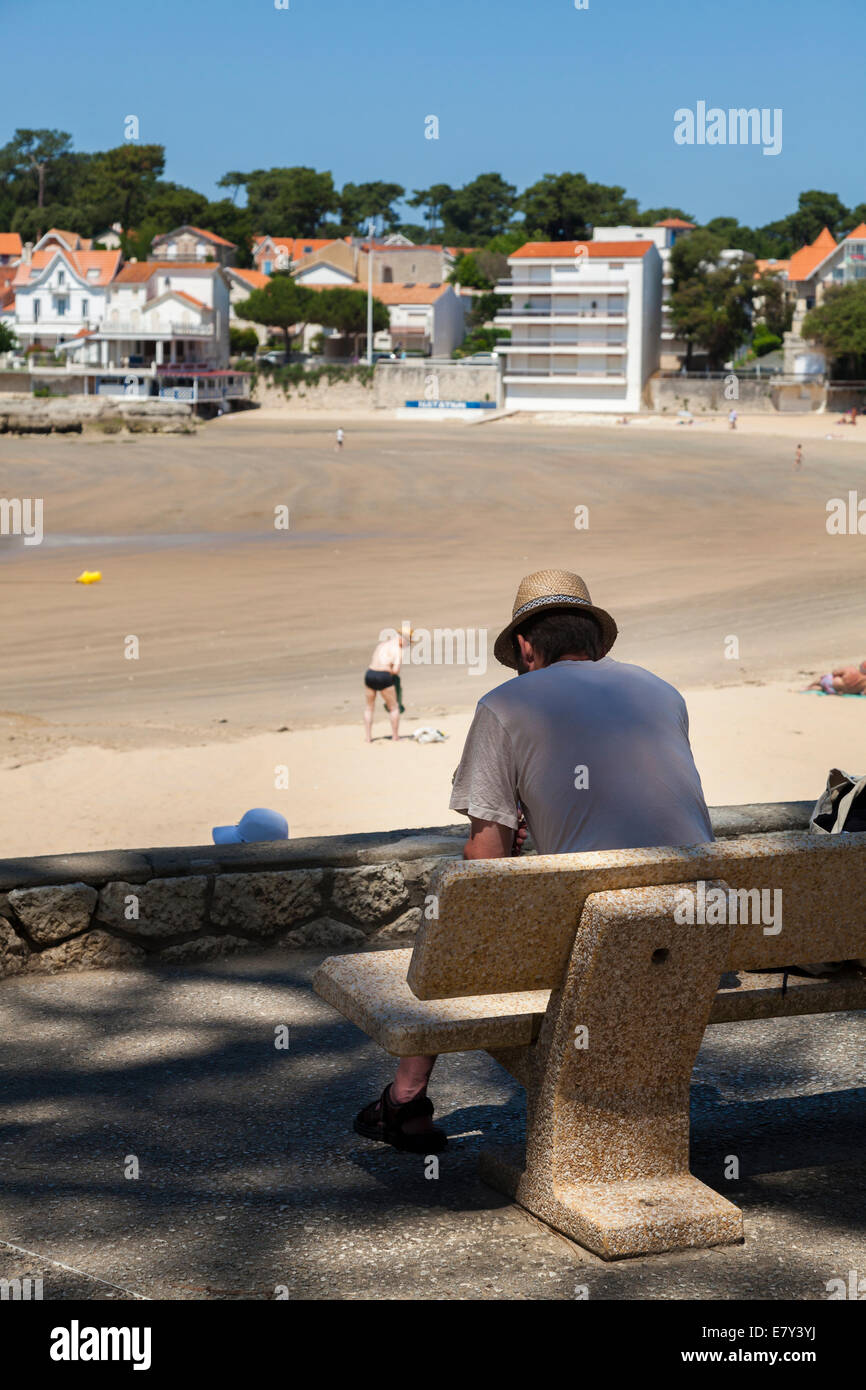 Homme assis sur un banc ombragé donnant sur la lecture de la plage d'adresses. Banque D'Images