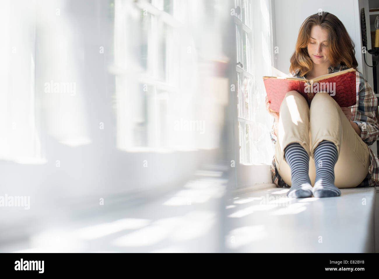 Une femme assise par une fenêtre, la lecture avec un livre sur ses genoux. Banque D'Images