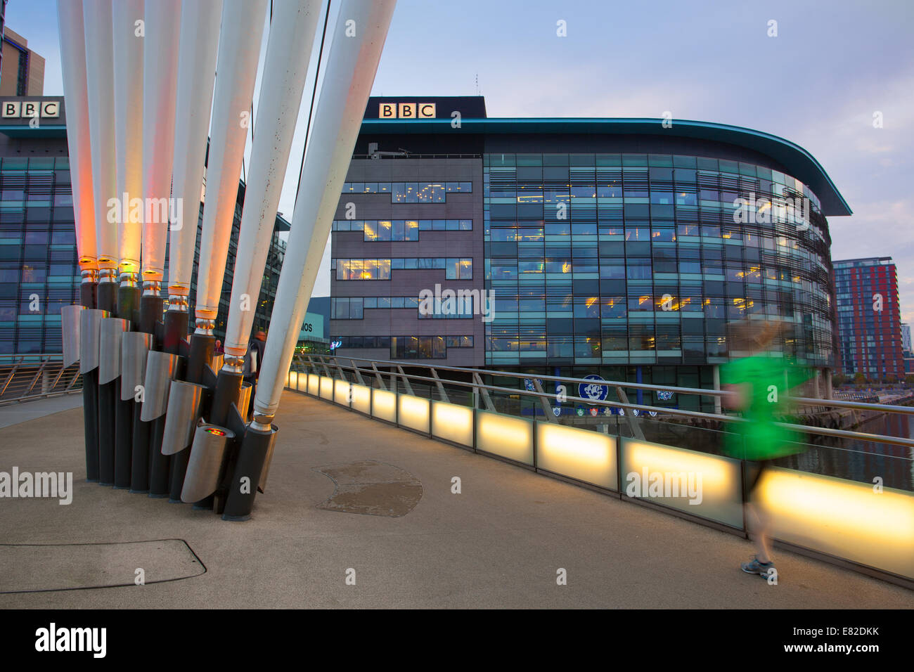 Le siège phare de la BBC dans le nord de la télévision de radiodiffusion à MediaCity, Media City, Salford Quays avec un jogger la passerelle de MediaCityUK illuminée à l'aube. Banque D'Images