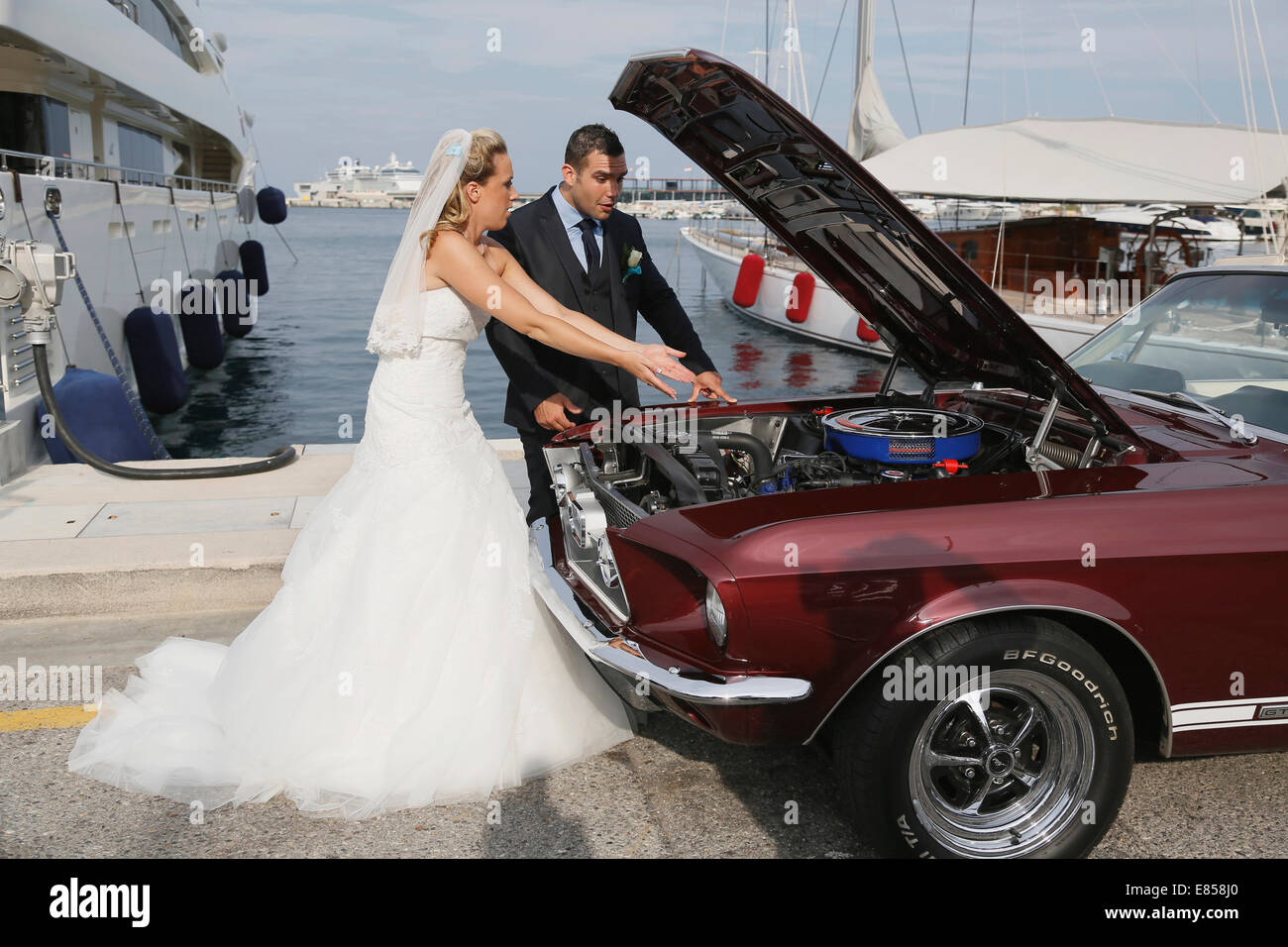 Mariée et le marié à la recherche dans le compartiment moteur d'une Ford Mustang après une panne, dans un port Banque D'Images