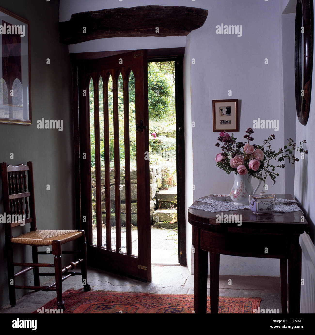 Vase de fleurs sur table console antique de cottage hall avec rush assis chaise à côté de la moitié porte ouverte sur le jardin Banque D'Images