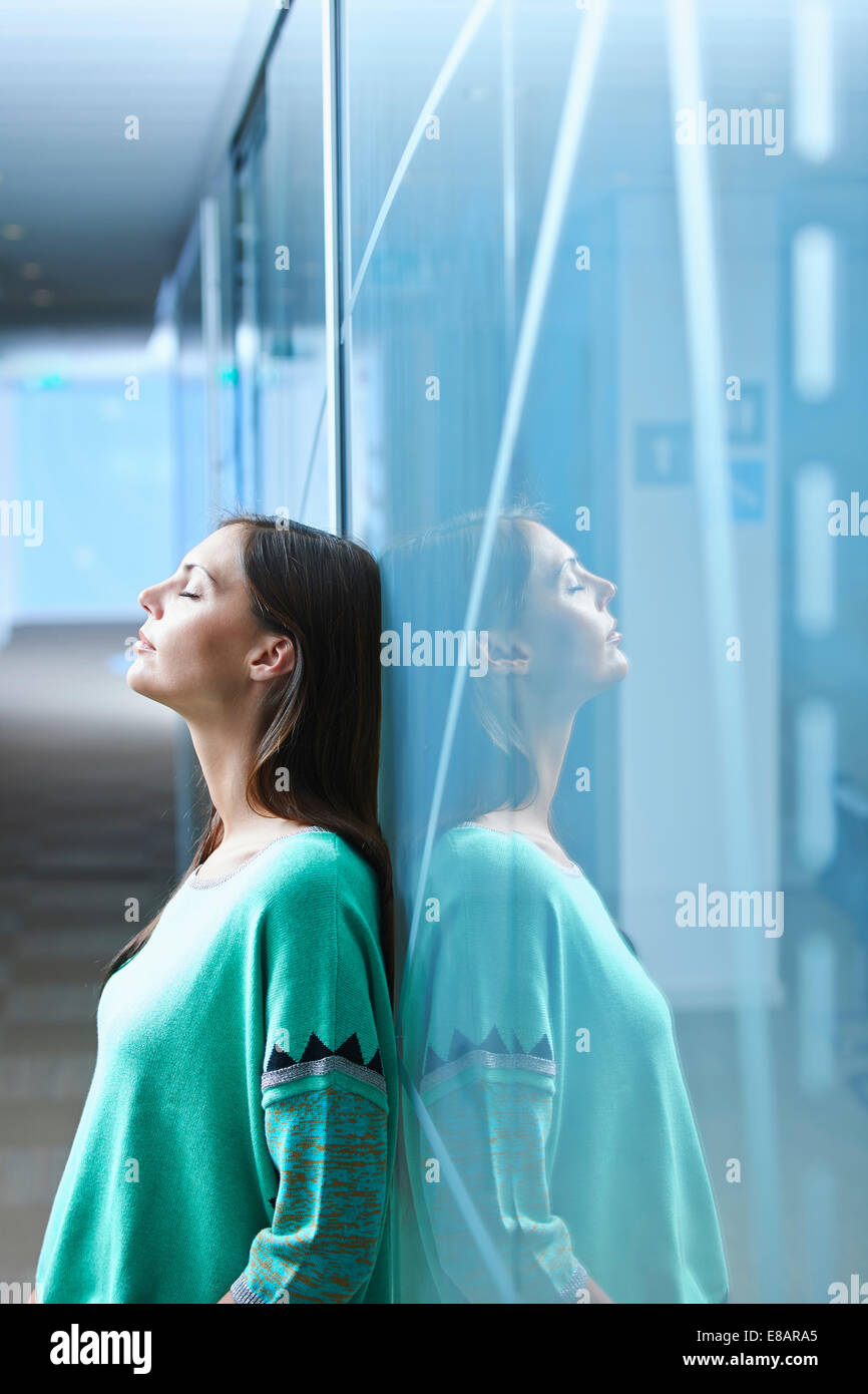 Mid adult businesswoman leaning against glass wall in office avec les yeux fermé Banque D'Images