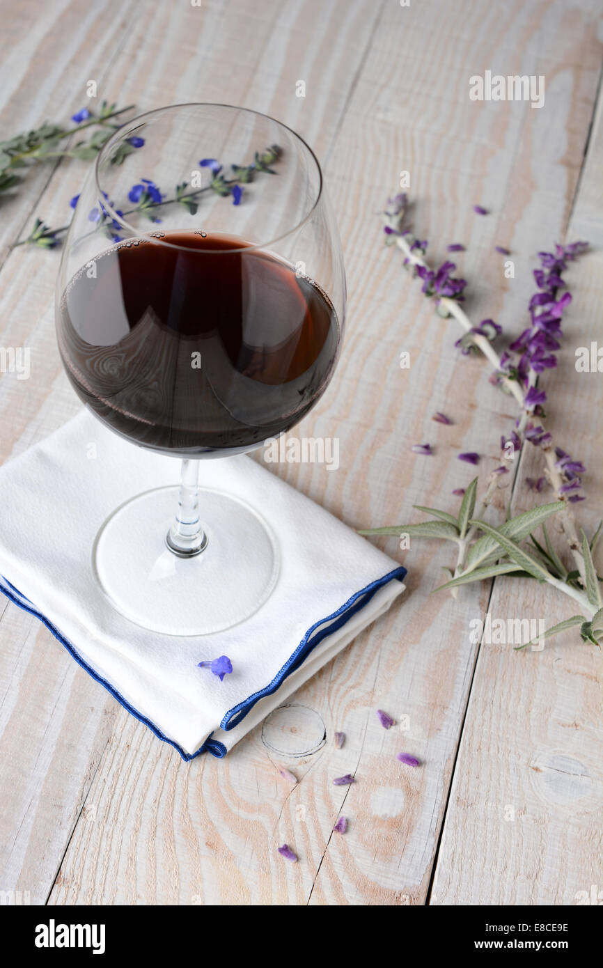 Un verre de vin rouge sur une serviette et une table rustique en bois blanc. Les fleurs et les pétales sont éparpillés sur la table. Format vertical Banque D'Images