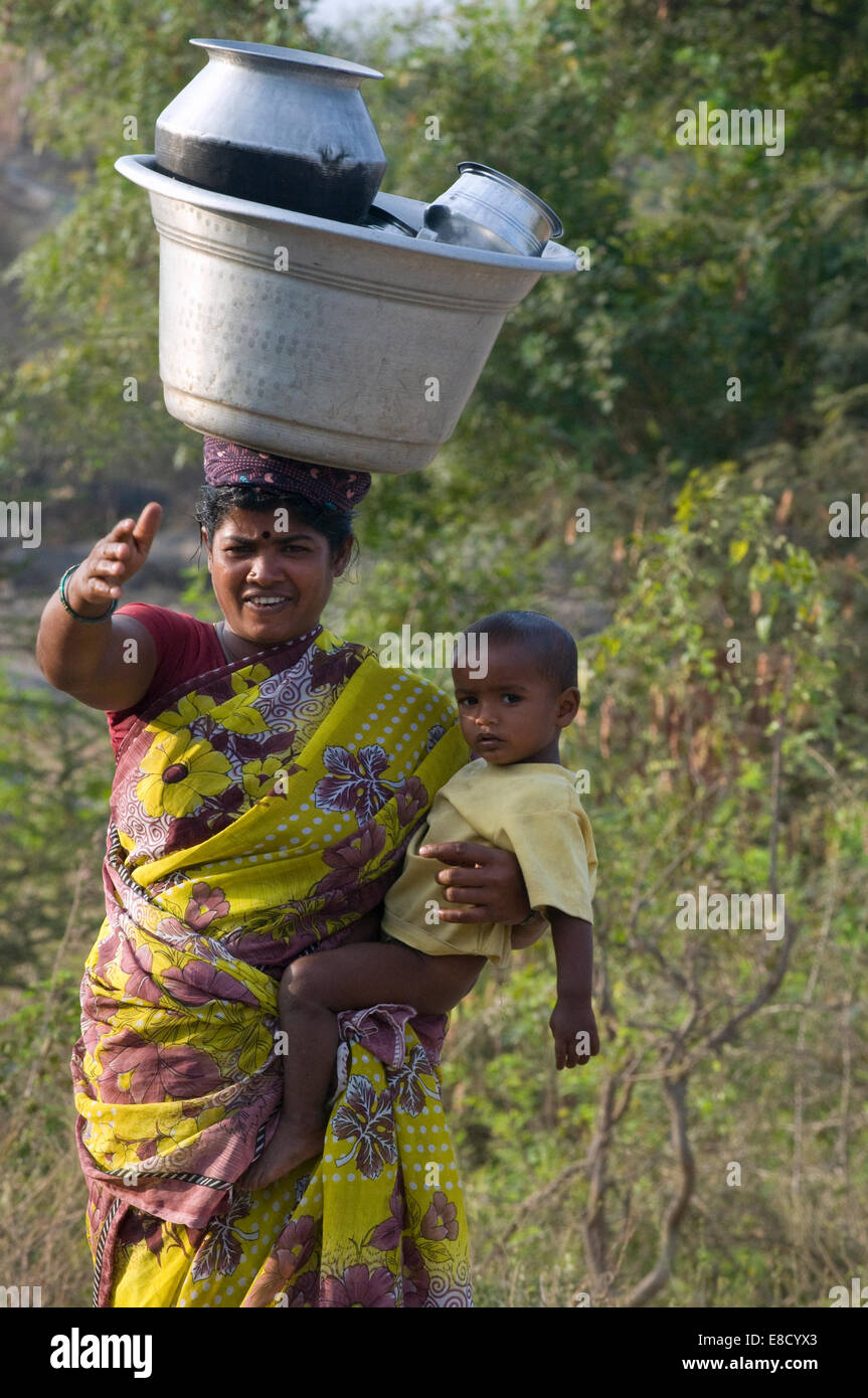 Mère indienne robuste habillé en sari coloré avec l'enfant sur la hanche avec cuisine casseroles et lave-bol en équilibre sur sa tête Banque D'Images