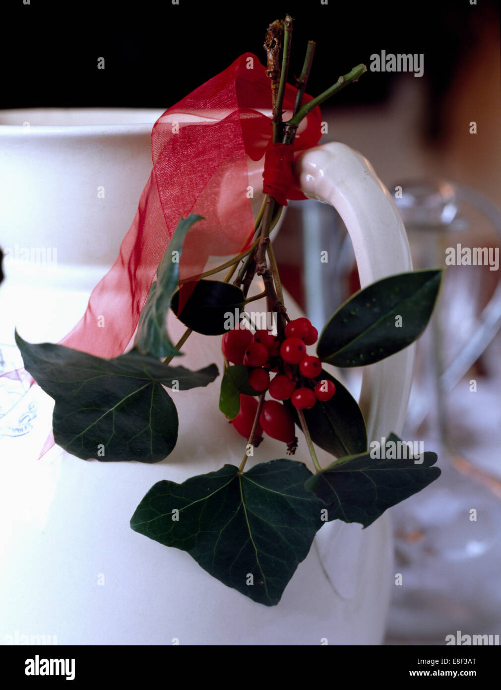 Close-up of a white ceramic jug décorées avec des baies de houx et de feuilles de lierre attaché avec un ruban organza rouge Banque D'Images