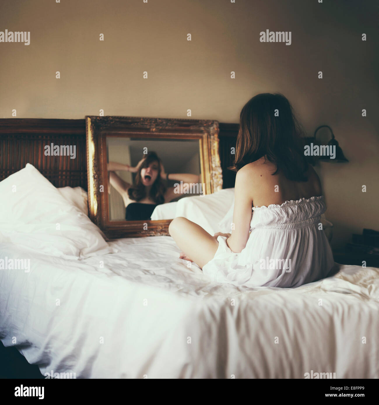 Femme assise sur le lit regardant dans le miroir regardant son reflet hurlant Banque D'Images