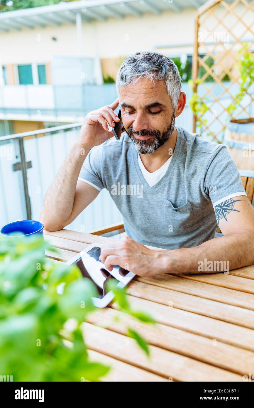 Portrait de l'homme assis sur son balcon téléphoner avec son smartphone lors de l'utilisation de digital tablet Banque D'Images