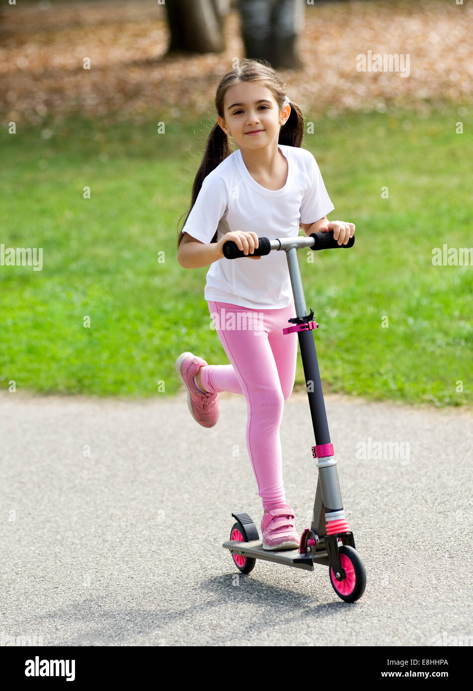 Jeune Fille en rose avec ses cheveux en amorces jouant sur un push scooter en plein air sur une route s'amusant et souriant Banque D'Images