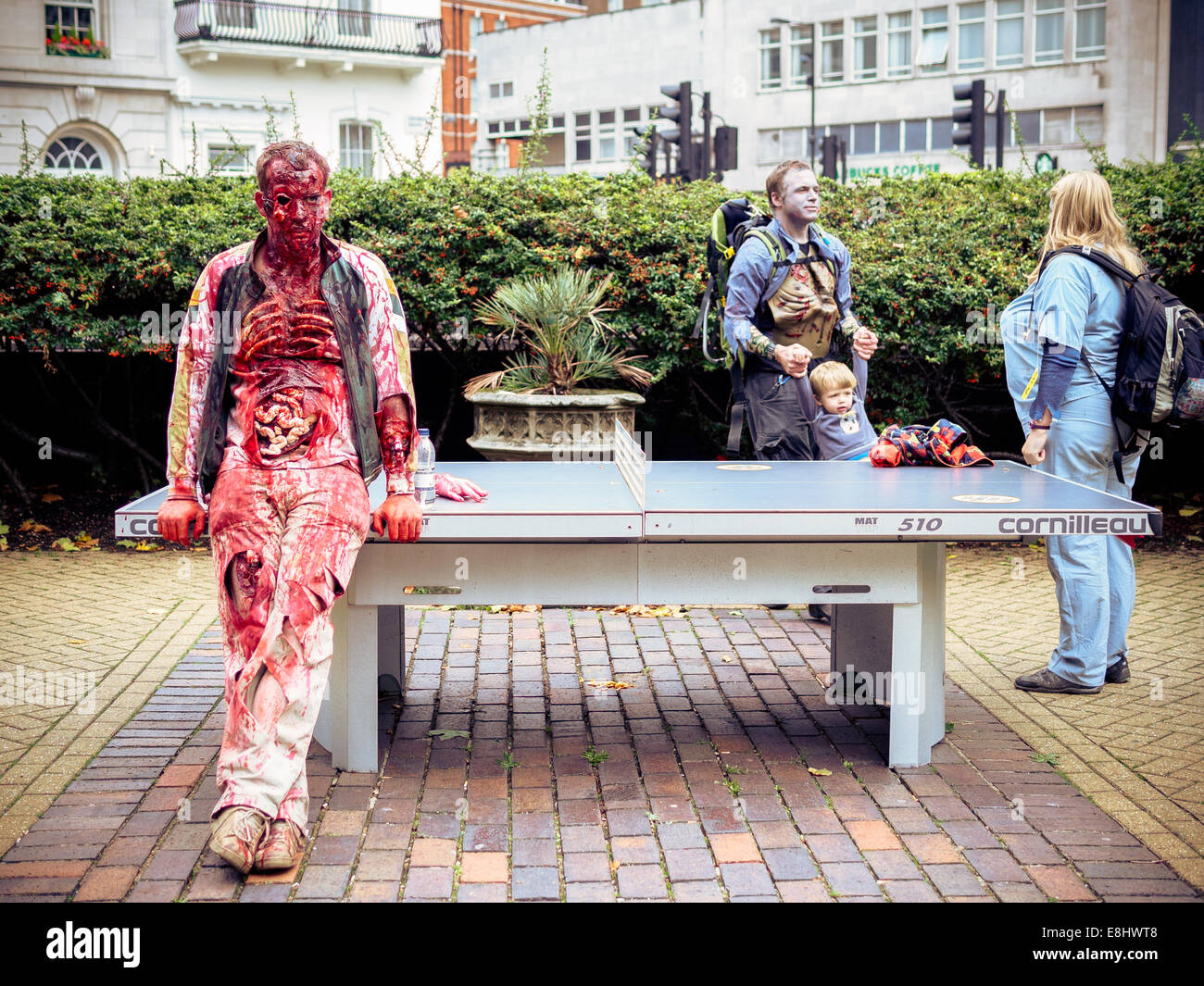 Zombie appuyé contre table de tennis de table, avec la famille zombie dans l'arrière-plan. Banque D'Images