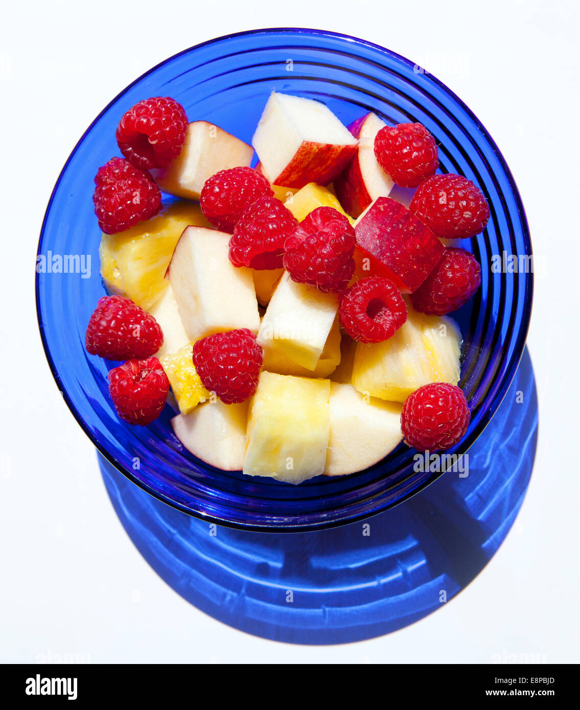Vue aérienne de framboises, de morceaux de pomme et d'ananas dans un bol en verre bleu cobalt against white background Banque D'Images