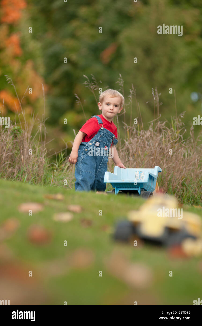 Une blonde deux-année-vieux garçon en salopette en jean joue avec des camions de jouets dans sa cour pendant l'automne. Banque D'Images