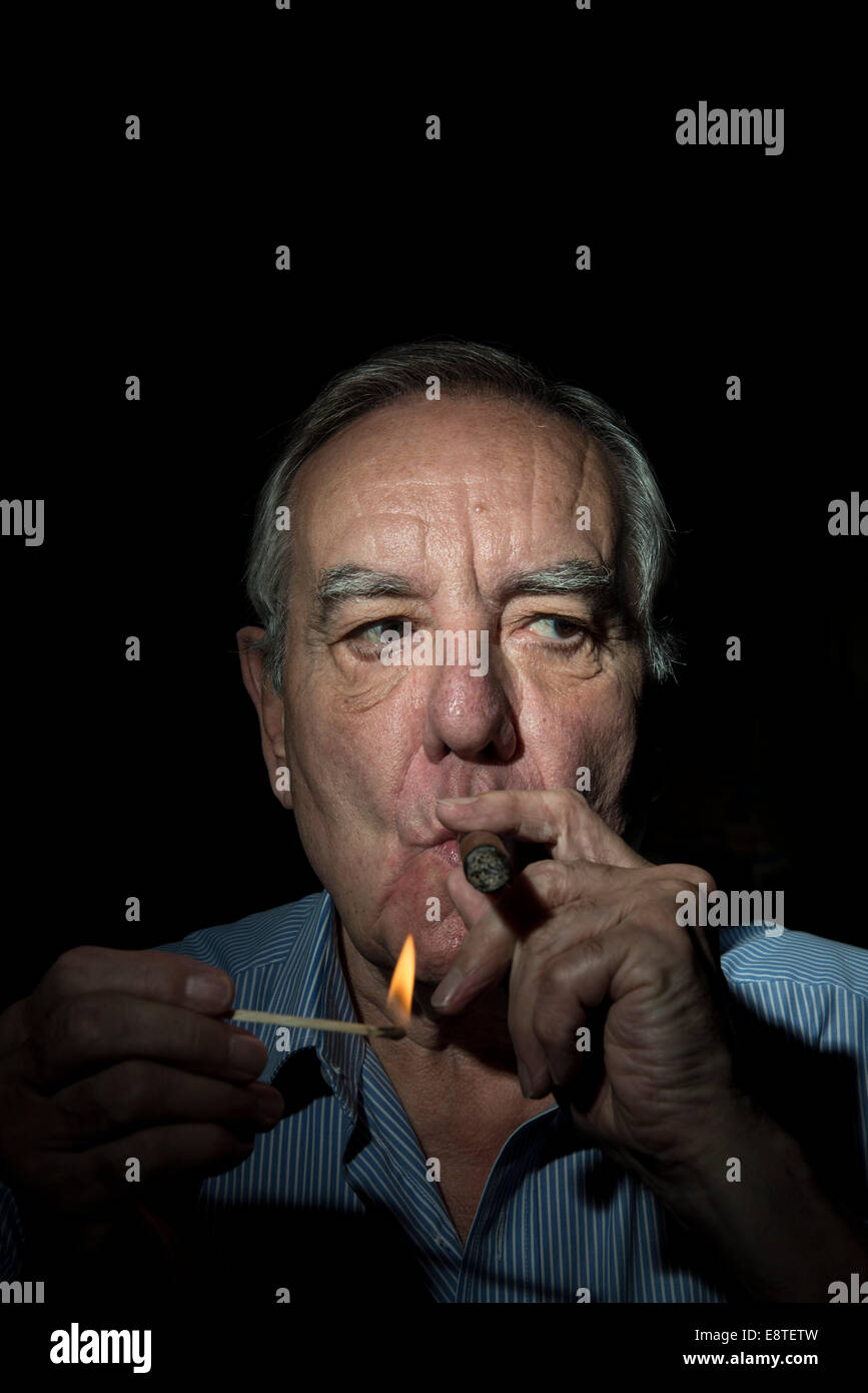 Un vieil homme blanc avec un visage de caractère, lumières et fume un cigare sur un fond noir, avec une photo de l'atmosphère sombre et Banque D'Images