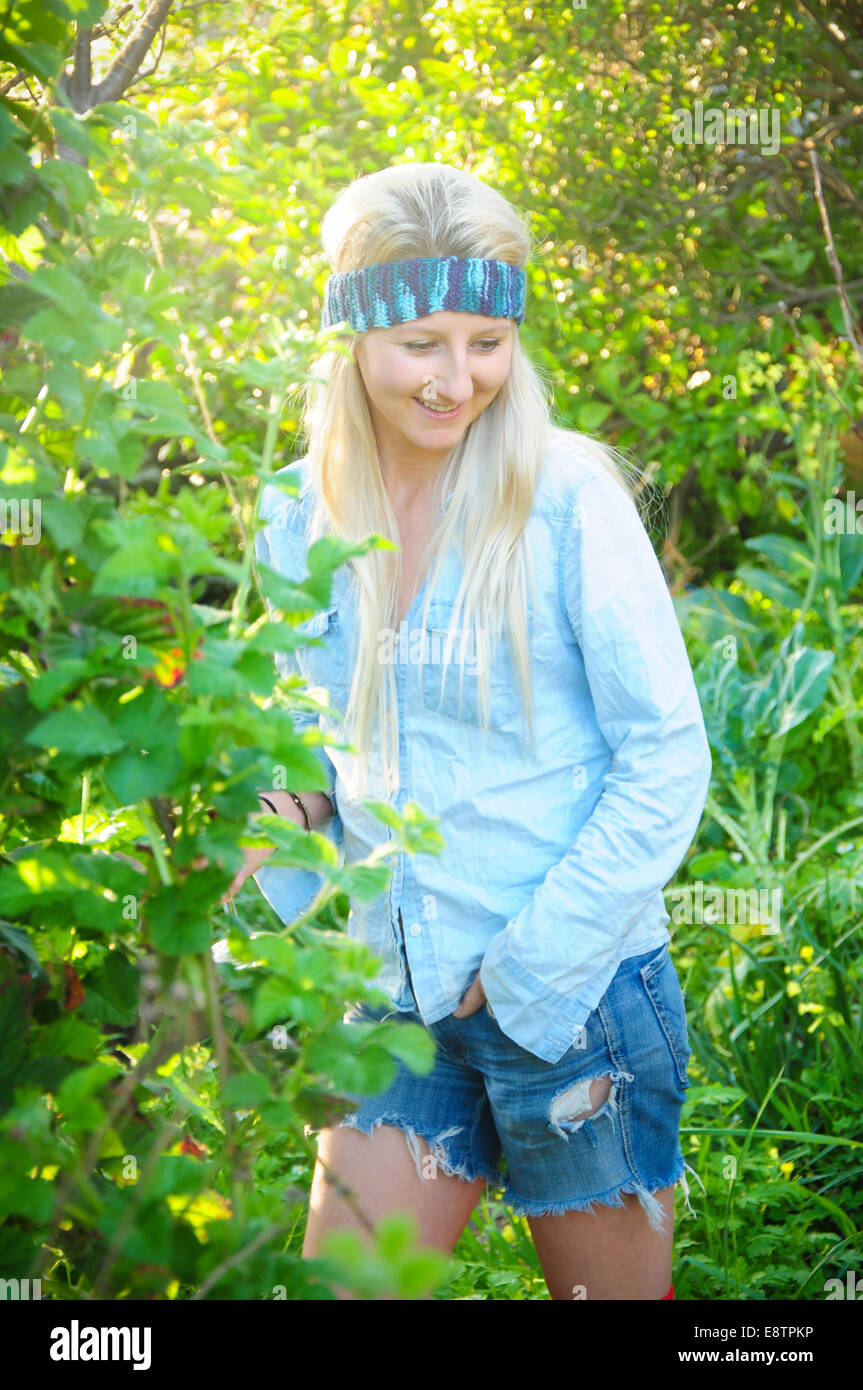 Une fille blonde se trouve dans un jardin avec sa main dans sa poche à la recherche vers le bas smiling portant un serre-tête fait maison et déchiré les shorts. Banque D'Images