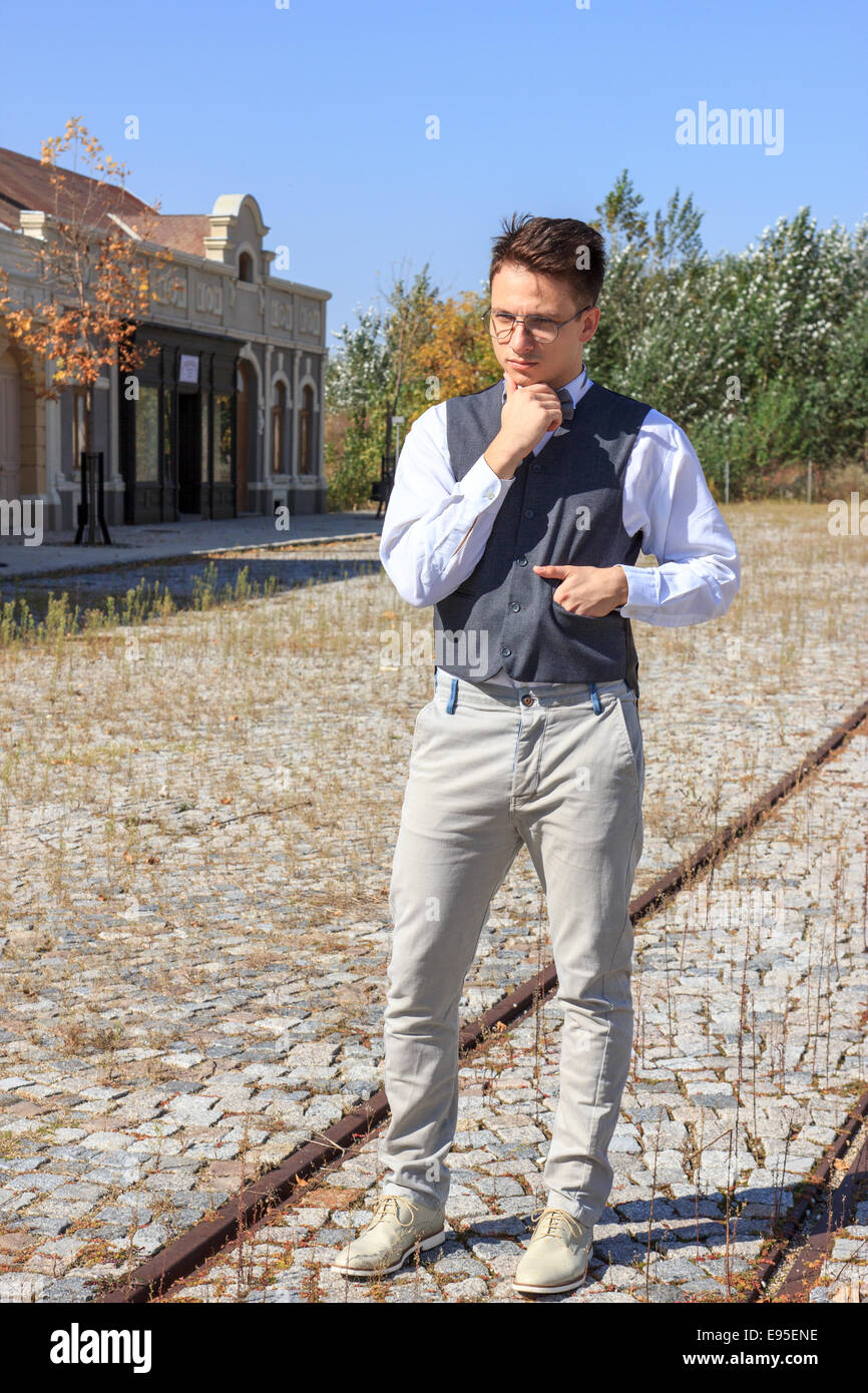 L'homme en chemise et gilet avec nœud papillon et des lunettes, debout dans des lignes de chemin de fer dans l'ancien vintage park ou ville et à côté Banque D'Images