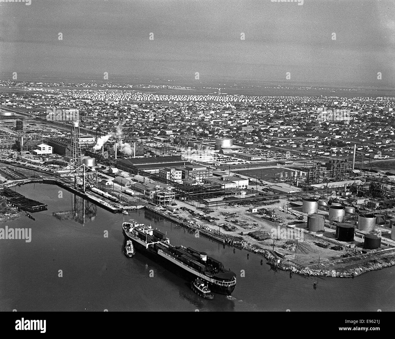 Une vue aérienne capture le Seatrain Louisiana, un cargo amarré à Texas City, avec une visibilité bien visible des opérations de Seatrain Lines. La scène comprend des barges, des cargos, des paysages urbains portuaires, des machines de levage, des wagons de chemin de fer, et les opérations de fret, mettant en évidence la logistique complexe des réseaux de la ceinture de coton et de l'ATSF Railway. L'image présente les principales activités de transport maritime, ferroviaire et remorqueur dans la zone du port industriel. Banque D'Images
