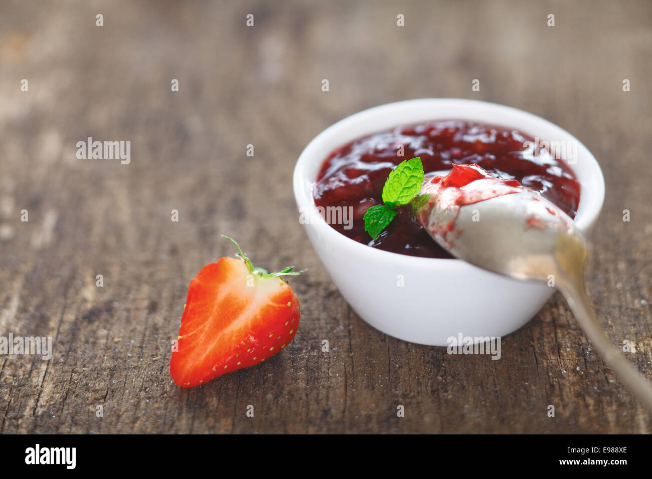 Petit plat de délicieux de confiture de fraises avec une cuillère à dessert d'argent sale en équilibre sur le rebord à côté d'une moitié rouge frais mûrs Banque D'Images