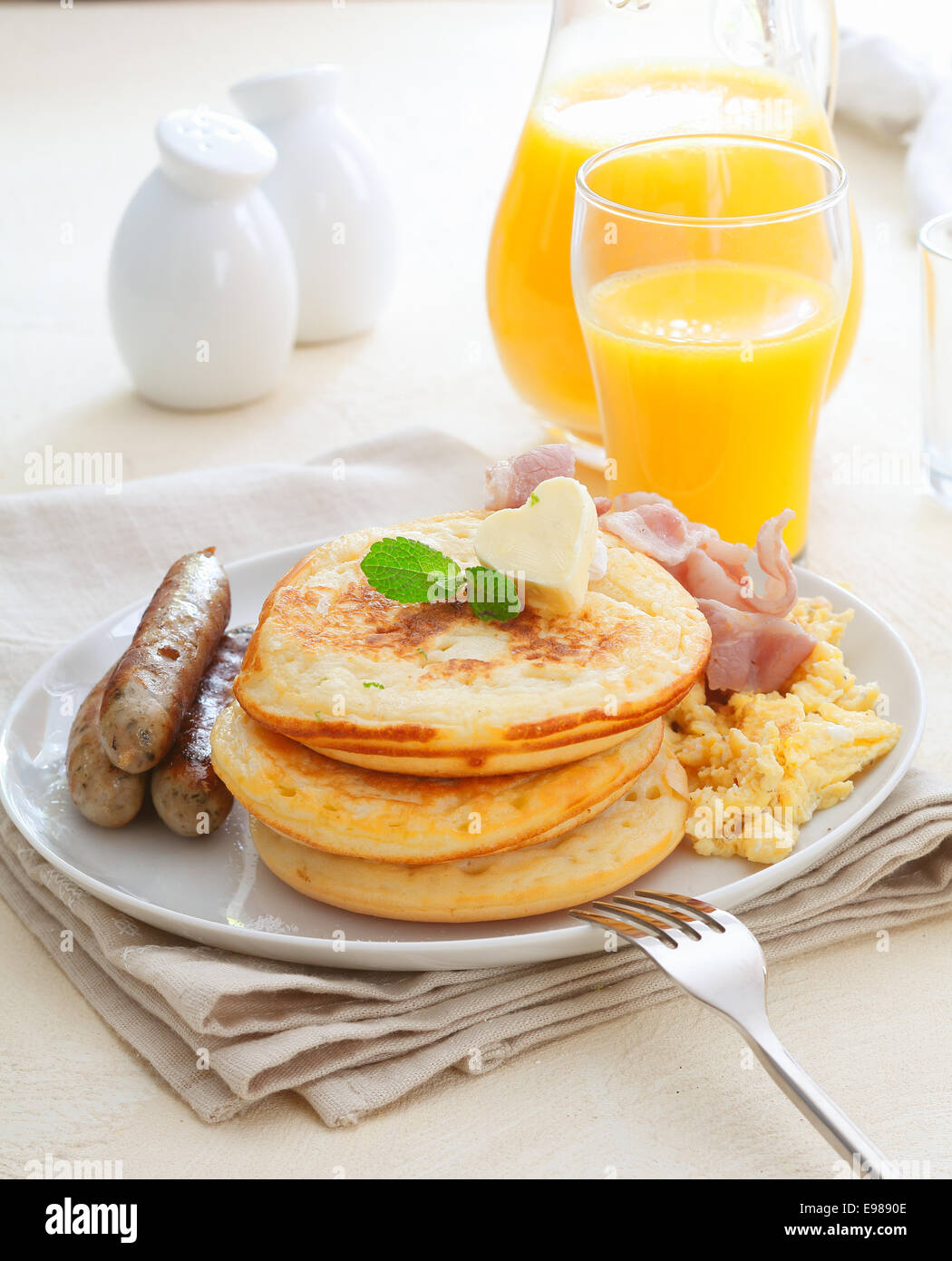 Petit-déjeuner nutritif sain avec saucisses, œufs, bacon et une pile de crêpes dorées avec un verre de jus d'orange sur une serviette Banque D'Images
