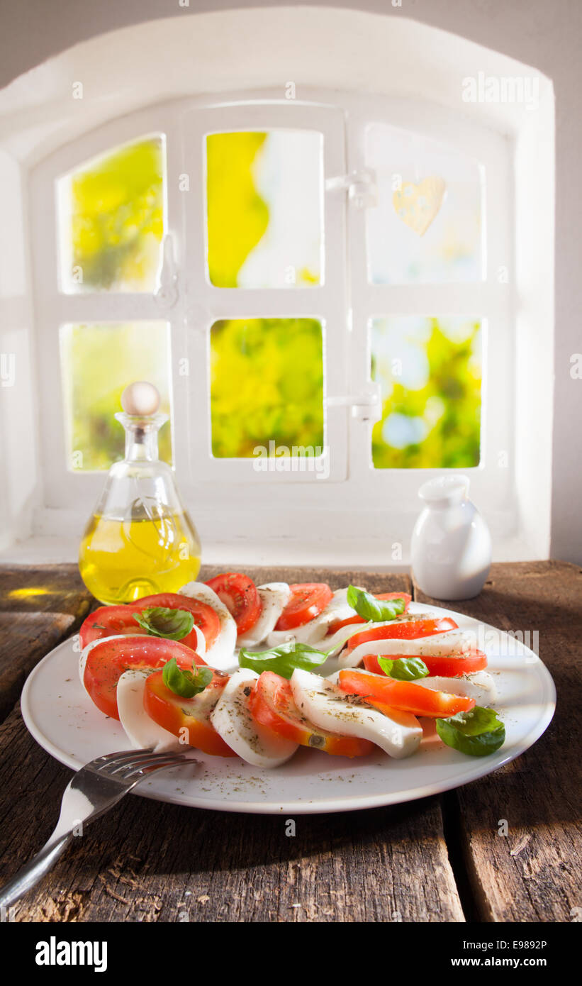 Servir sur une assiette pour d'autres tranches de tomate et de fromage salade avec vinaigrette à l'huile sur une table en bois près de la fenêtre d'un pays Banque D'Images
