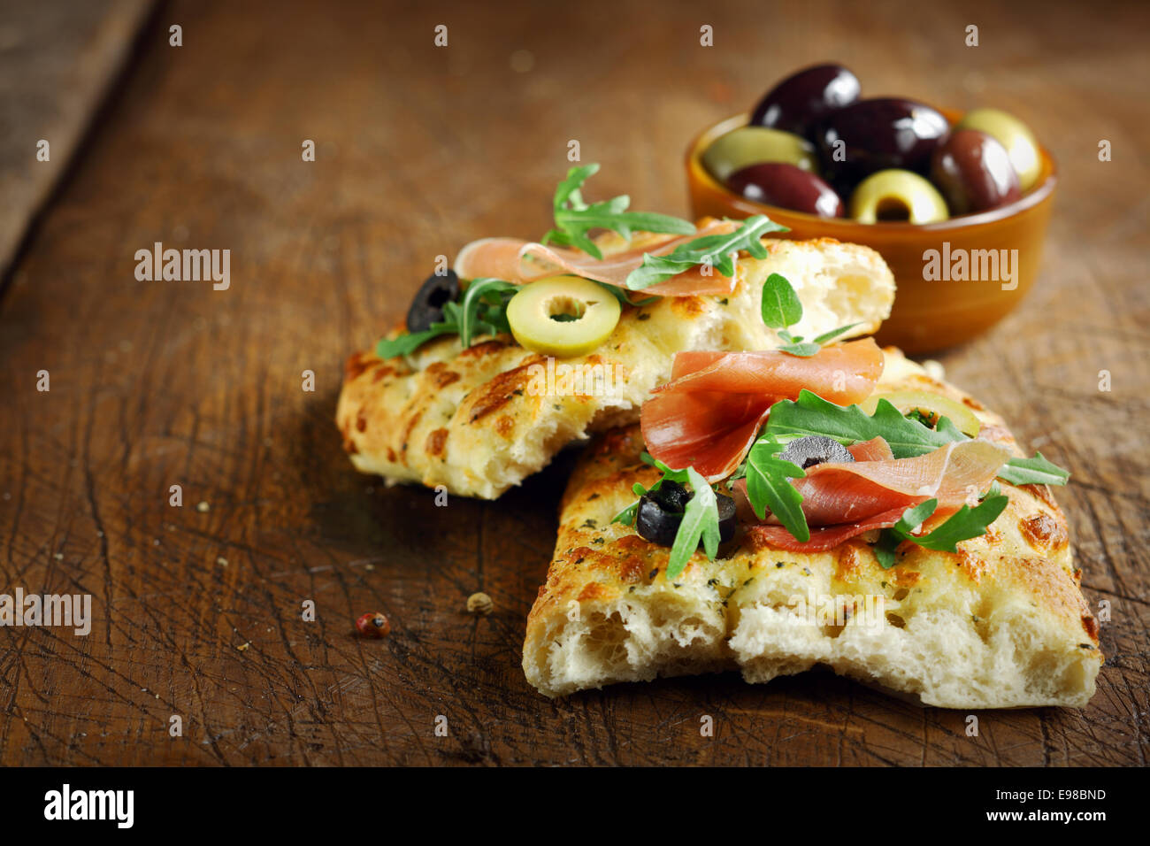 Jambon et à la roquette sur pain focaccia italienne fraîche garnie d'olives tranchées allongé sur une vieille table de cuisine en bois haut Banque D'Images