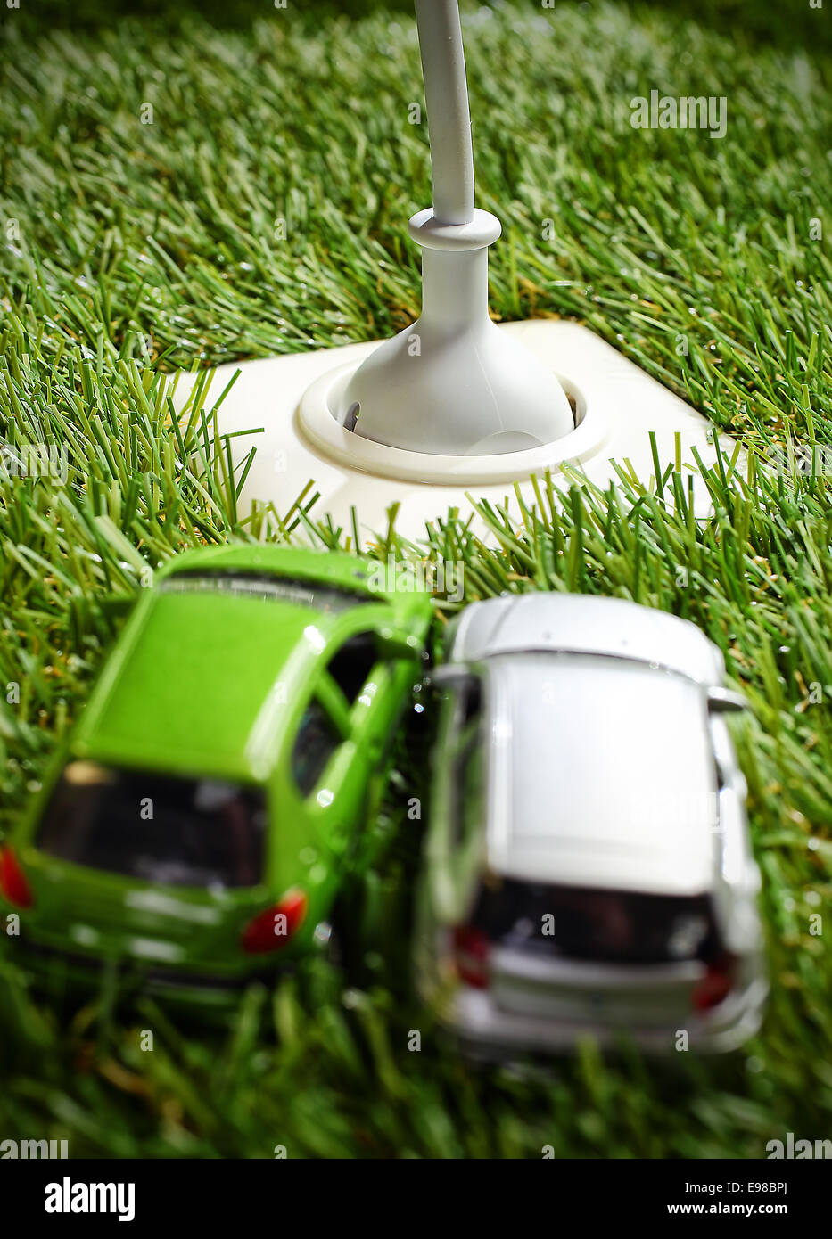 Prise d'alimentation électrique de plein air dans l'herbe verte avec un connecteur et câble inséré et deux modèles de voitures jouets en premier plan Banque D'Images
