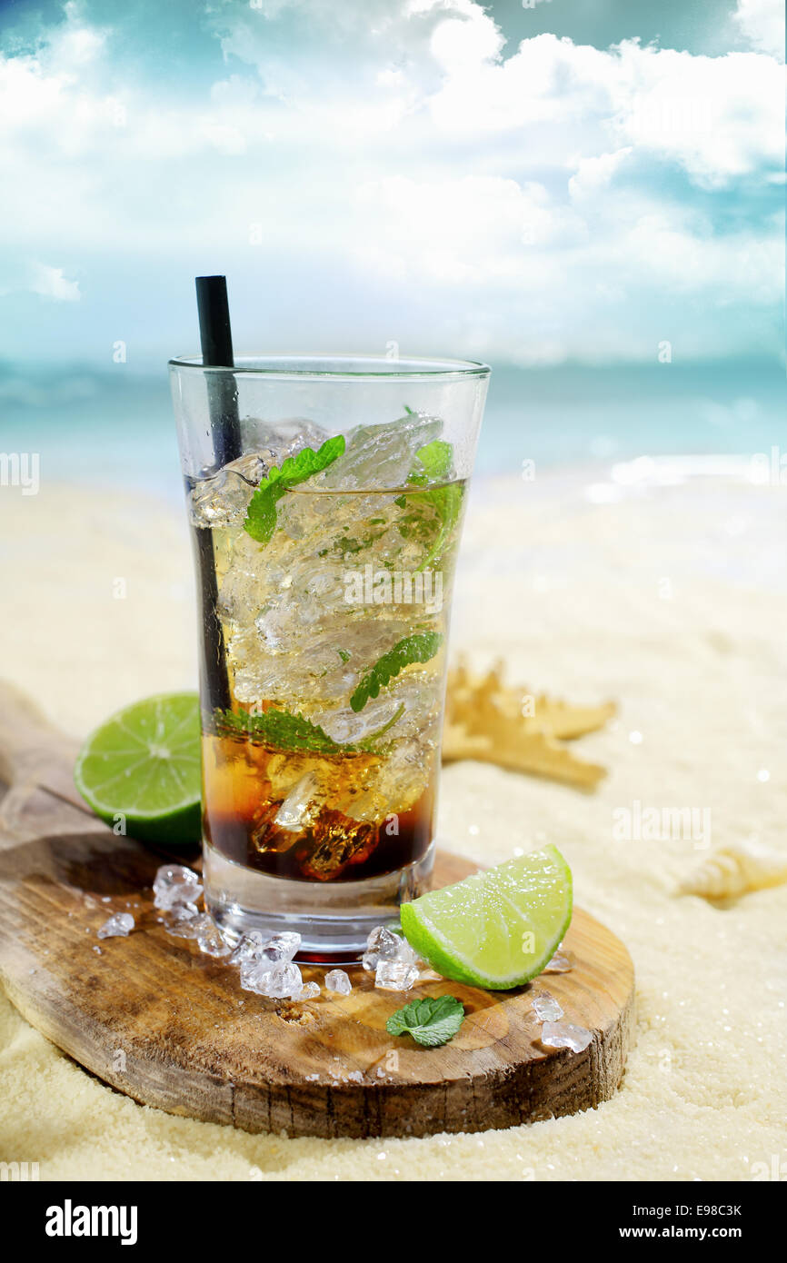 Cocktail au rhum glacé rafraîchissant servi dans un grand verre sur une planche en bois avec des tranches de citron sur le sable doré d'une plage tropicale idyllique Banque D'Images