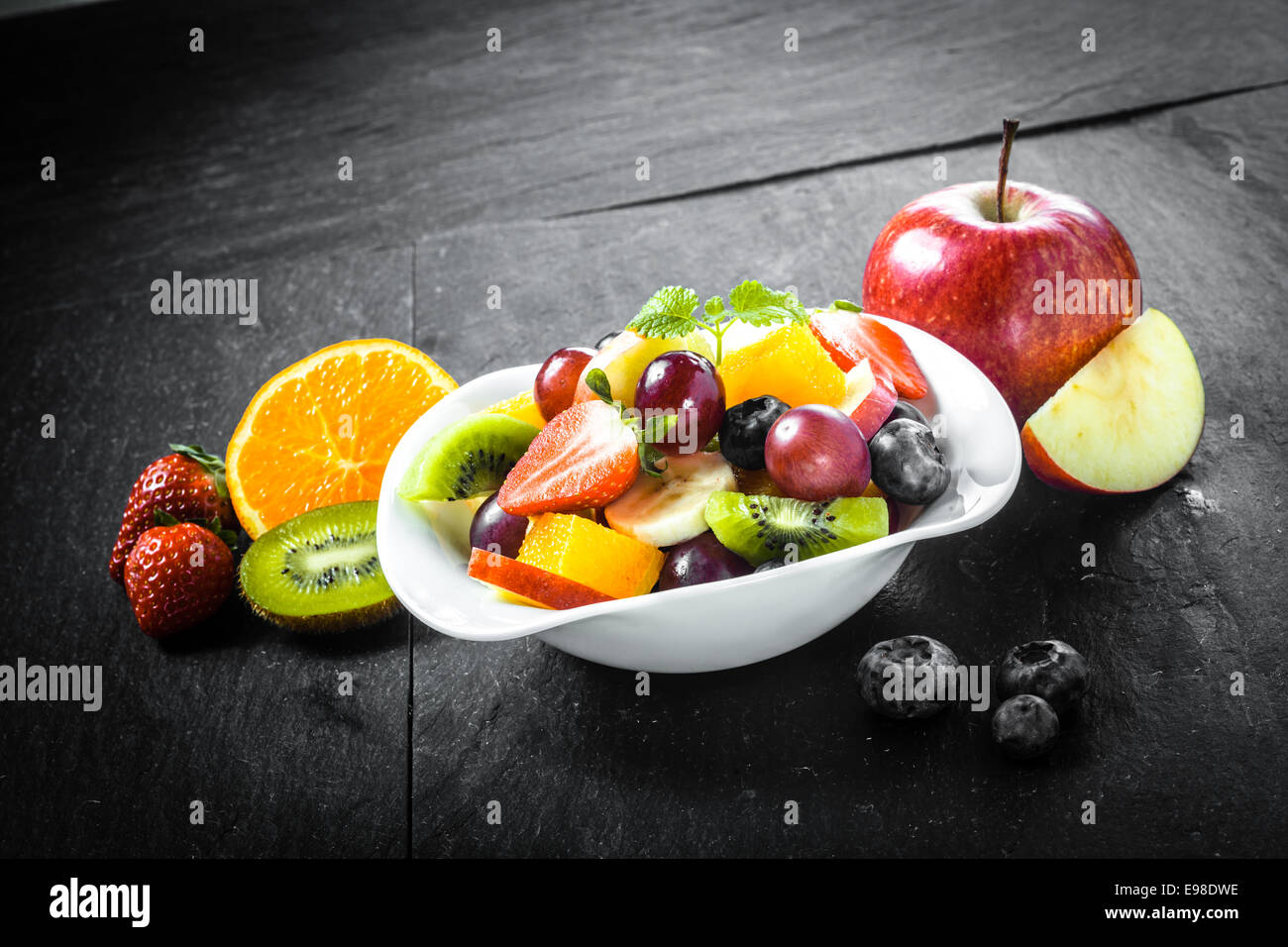 La préparation d'un délicieux bol de fruits frais colorés avec salade de fruits tropicaux, y compris les bleuets, pomme, orange, les kiwis et les fraises sur une surface ardoise cuisine Banque D'Images