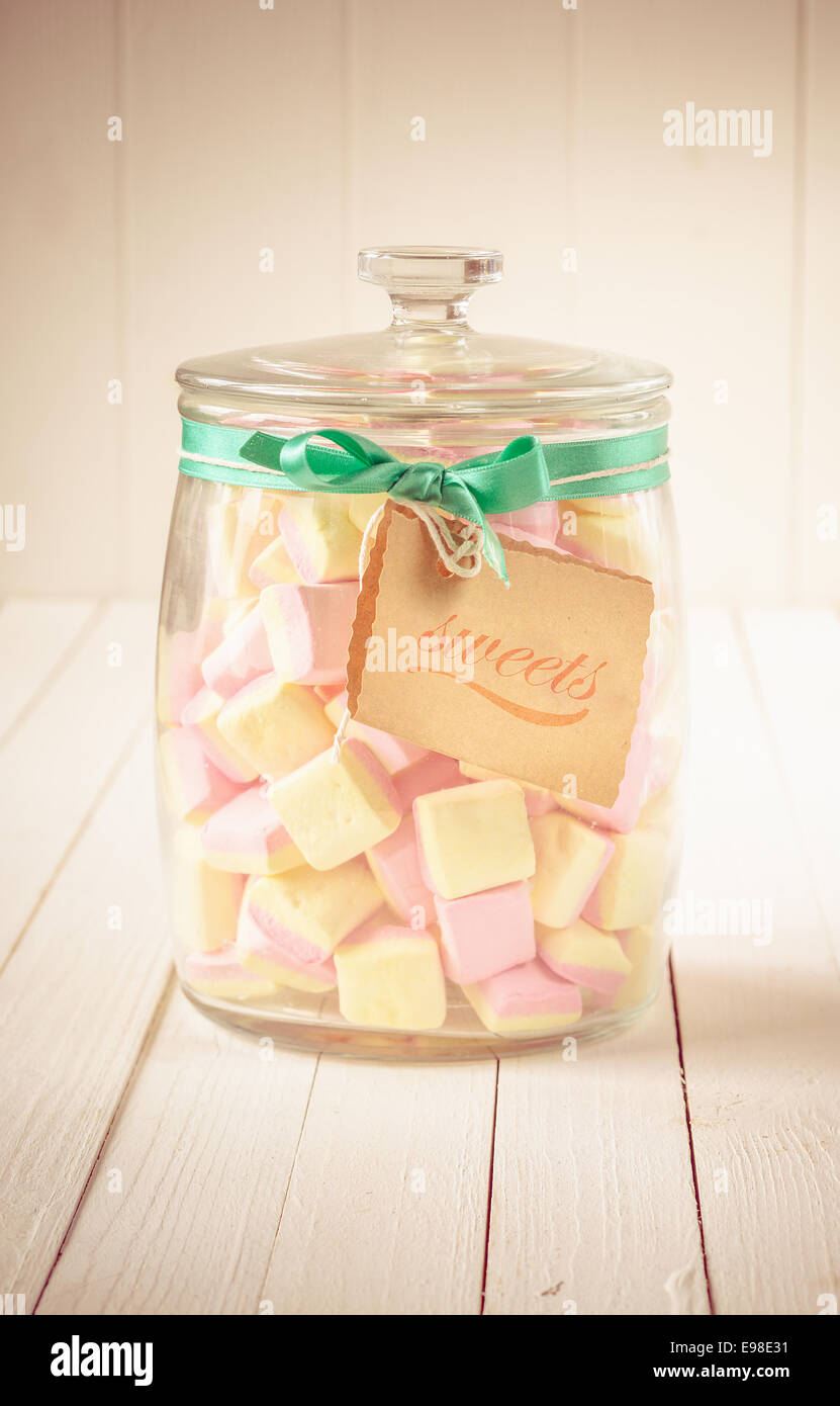 Verre ancien bocal rempli de bonbons carrés avec guimauves rose et jaune et une étiquette attaché avec un ruban vert sur un fond de bois blanc Banque D'Images