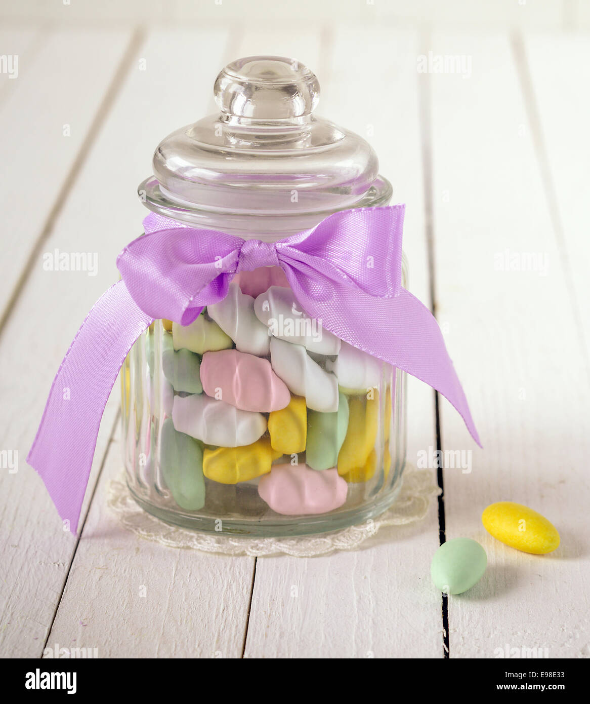 Gros plan d'un verre ancien bocal rempli de bonbons colorés avec Sucre et amandes un arc violet sur un fond de bois blanc Banque D'Images