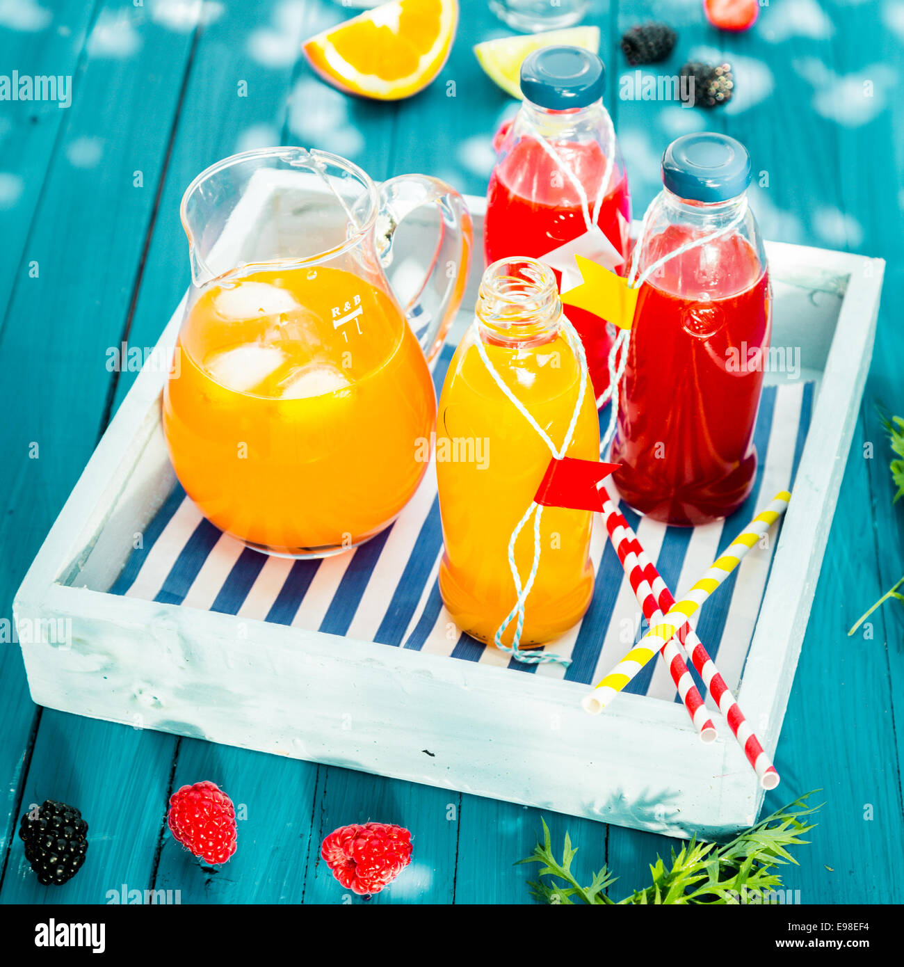 Des jus de fruits orange et servi dans des bouteilles en verre et réfrigérés avec de la glace dans une cruche en verre coloré bleu, sur une table de pique-nique en bois sur un plateau en bois Banque D'Images