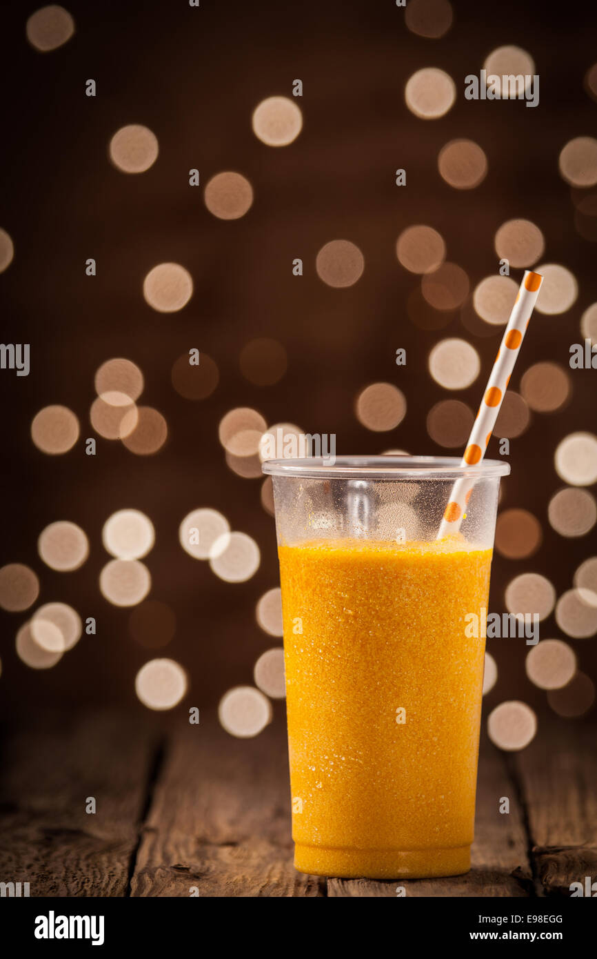 Verre de plastique orange tropicale glacée rafraîchissante savoureux Smoothie mangue debout sur un comptoir en bois donnant sur une partie de l'effet bokeh scintillant lumières avec copyspace Banque D'Images