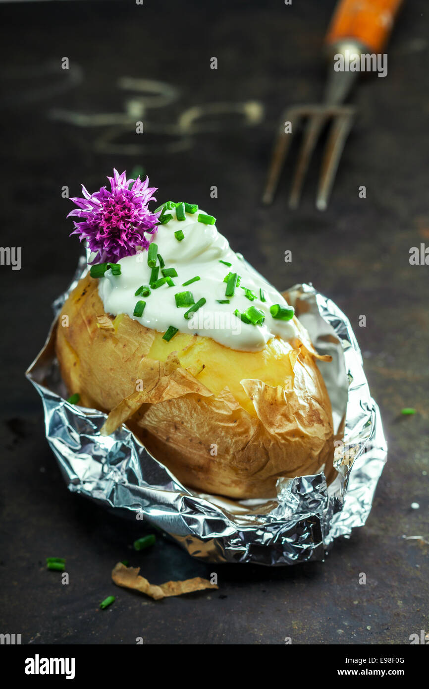 Pomme de terre au four aluminium jacket servi dans l'aluminium aluminium garnie de crème sure et de ciboulette fraîche, hachée et garni d'une fleur de ciboulette mauve Banque D'Images