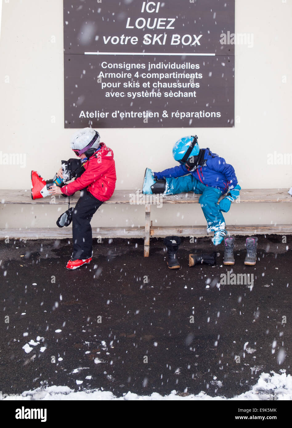 Petit garçon et fille en vêtements de ski casques et lunettes assis sur un banc, mettre sur les chaussures de ski avec la neige qui tombe Banque D'Images