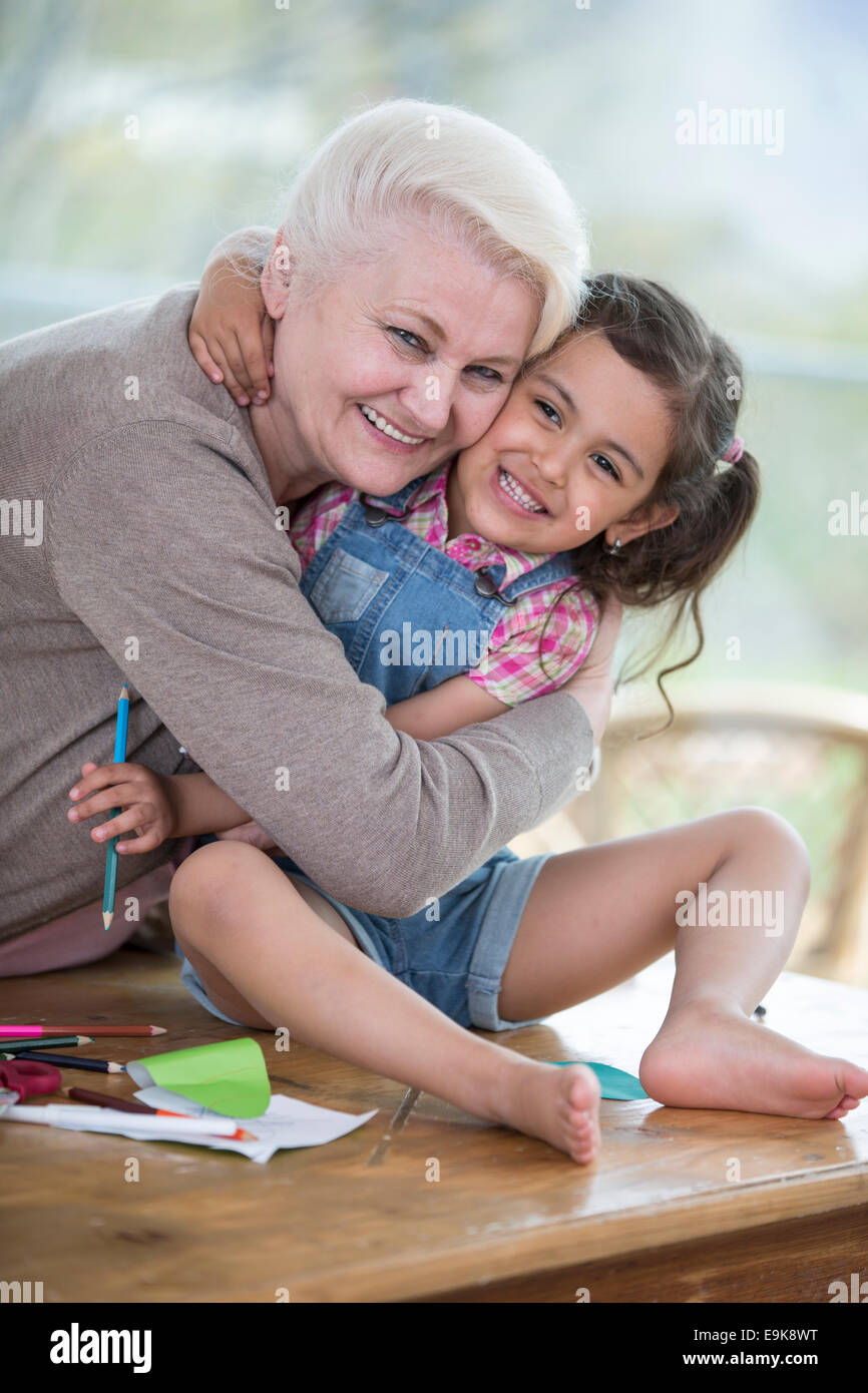 Portrait of senior woman hugging granddaughter sitting on table à la maison Banque D'Images