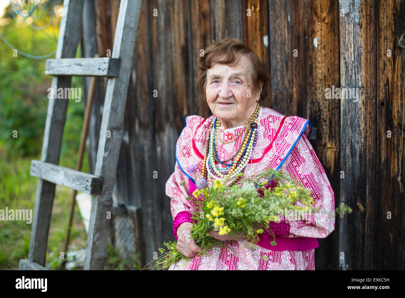 Vieille Femme dans vêtements ethniques avec des fleurs sauvages dans leurs mains dans la cour de sa maison. Banque D'Images