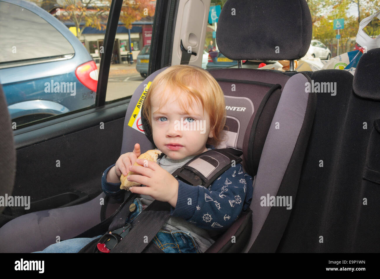 Un garçon âgé d'un an assis dans un siège de voiture dans une voiture en stationnement au parking d'un supermarché Banque D'Images