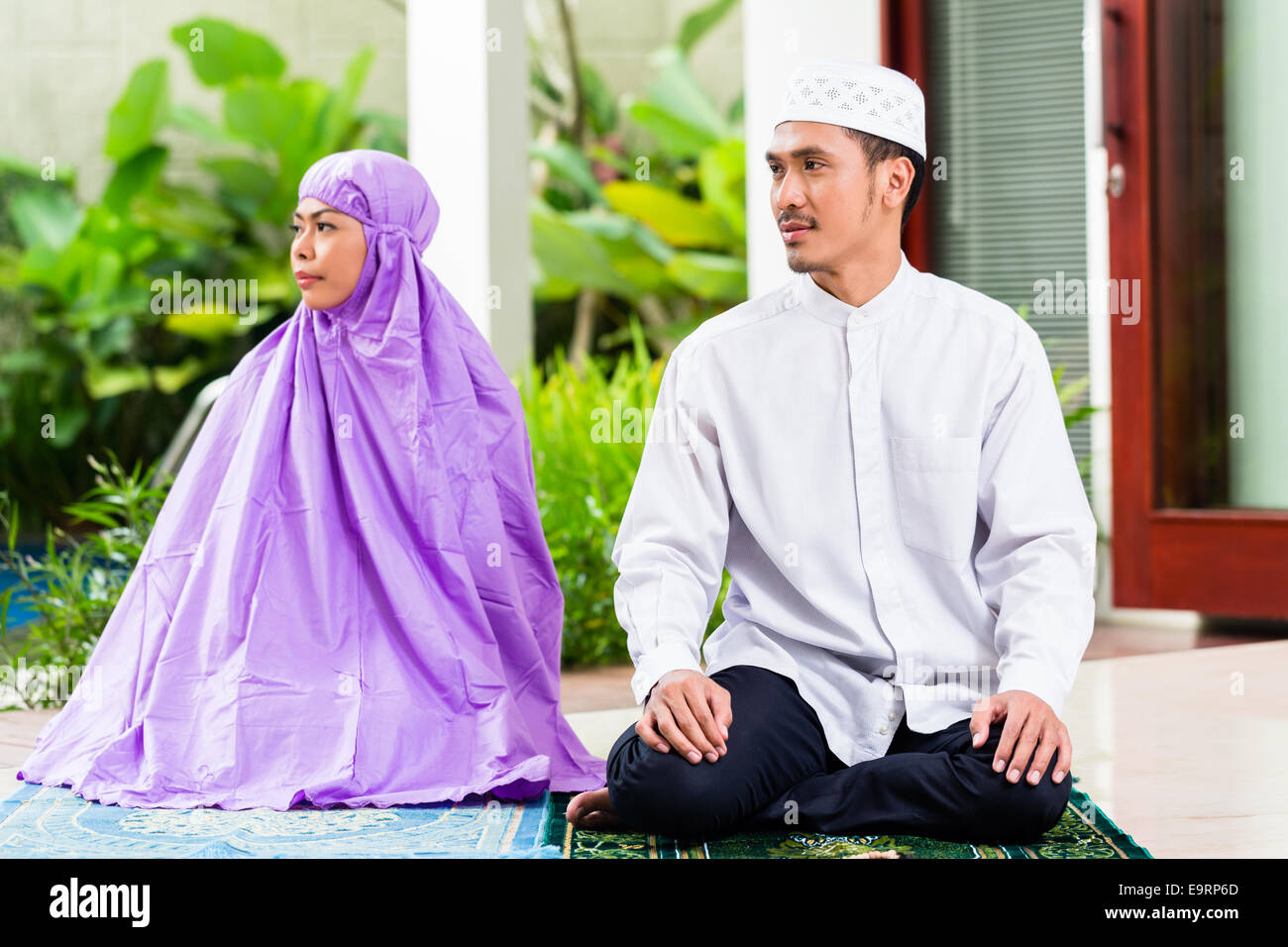 Couple musulman d'Asie, l'homme et de la femme, en priant à la maison assis sur les tapis de prière dans leur maison devant le jardin tropical Banque D'Images